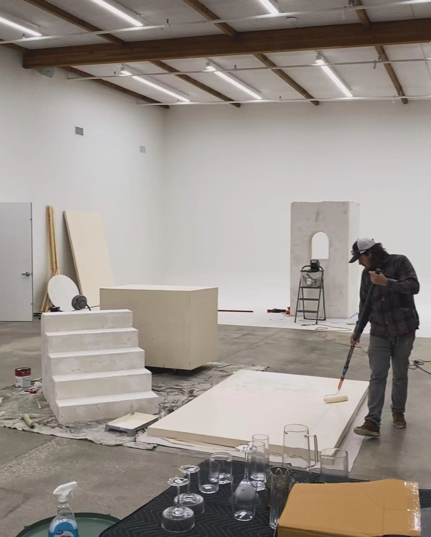 Interior of an art gallery or event space under construction with a person painting the floor. White walls, some art or display boards leaning against the wall, and a small set of stairs. The ceiling has exposed beams and lighting fixtures.