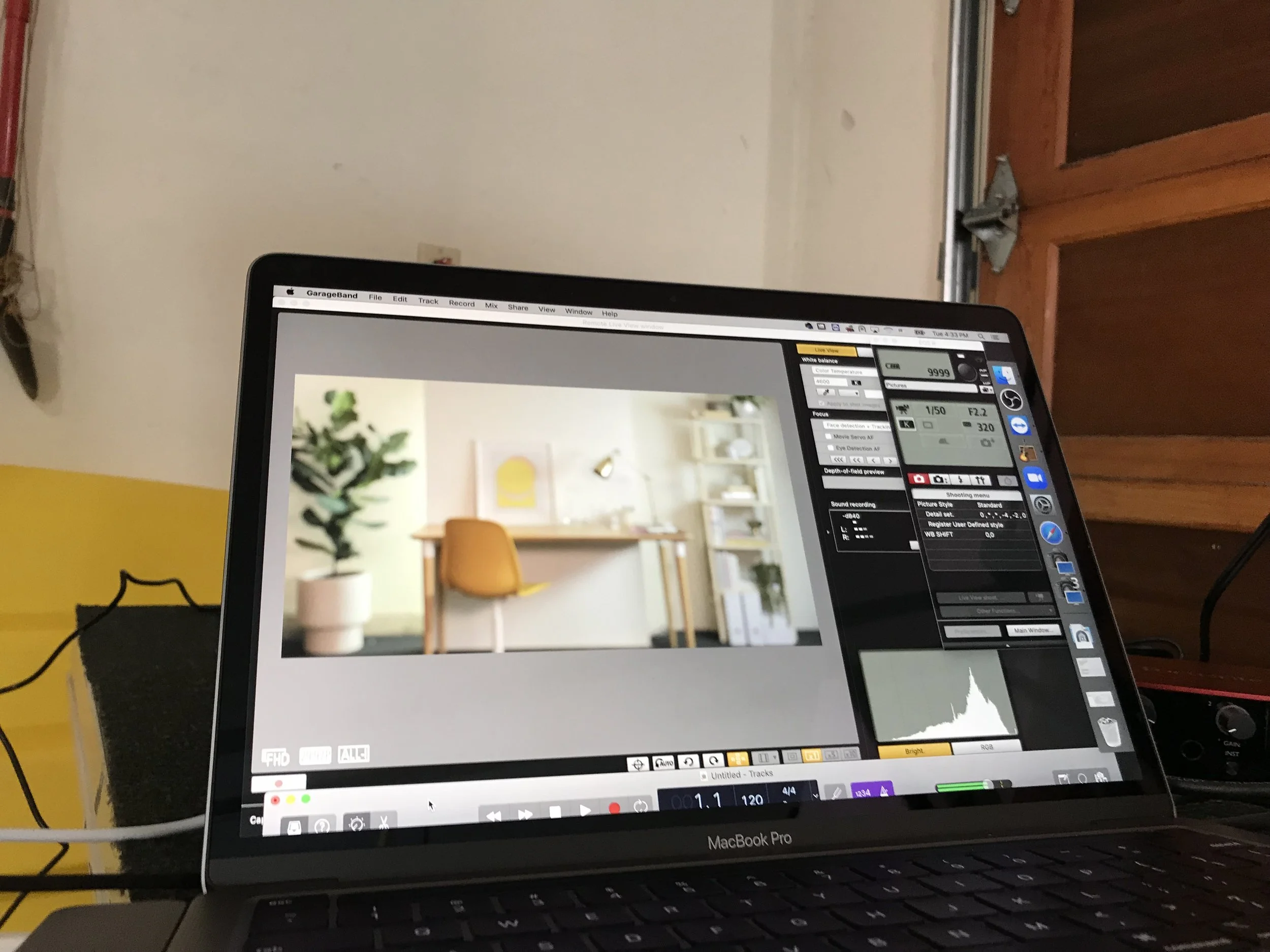 A MacBook Pro laptop displaying a photo editing or audio editing program, with a blurred background of a room with a yellow wall, shelf, and a chair.