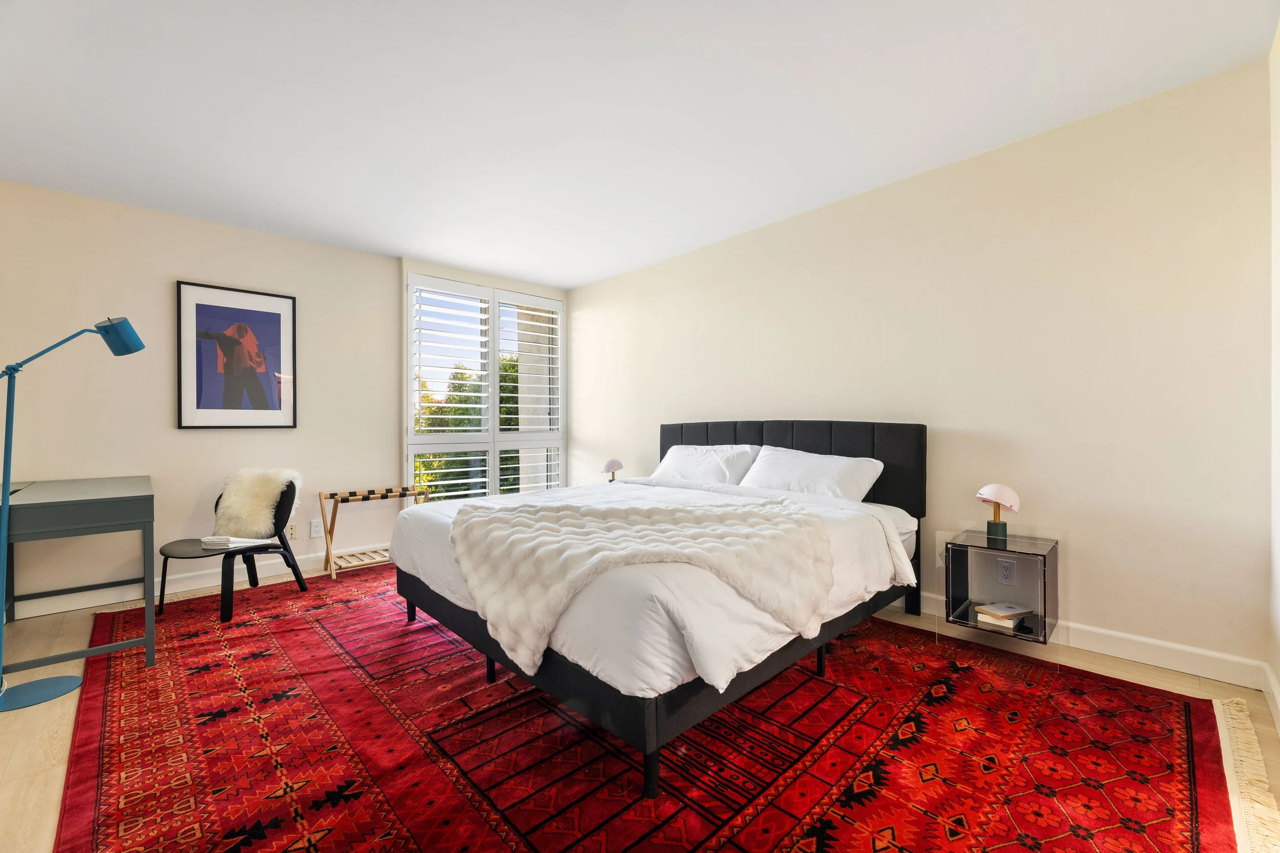 A bedroom with a large bed, white bedding, and a black headboard. There is a red patterned rug under the bed. To the left, a gray desk with a blue lamp and a black chair with white fur and papers on it. A framed picture above the desk depicts a person in front of a colorful background. Sunlight streams through the window with white shutters. There is a small bedside table with a pink lamp and a small black table with books on the right.