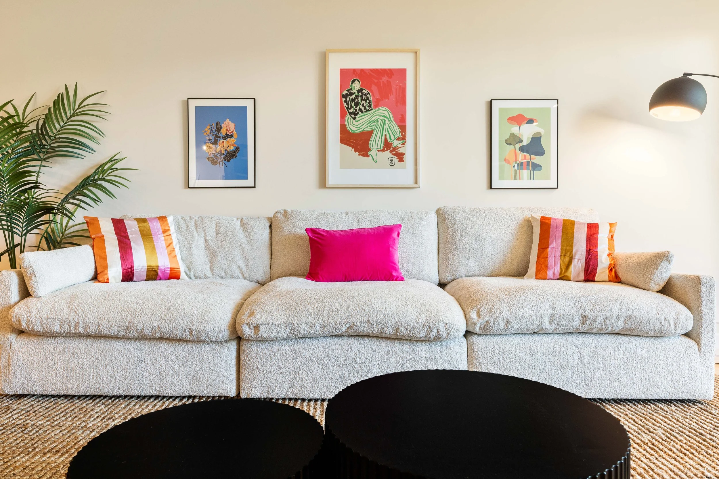 Living room with beige couch, colorful striped pillows, framed artwork on cream wall, green plant, black and beige rug, and black floor lamp.