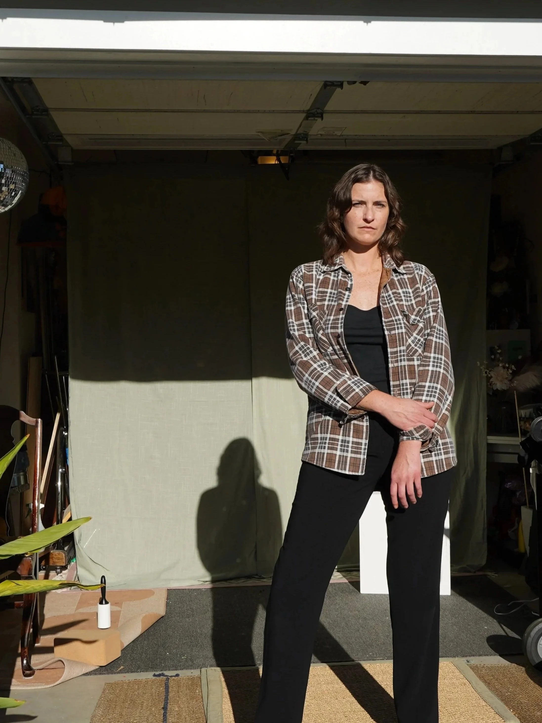 A woman standing inside a room with a dark background, wearing a plaid shirt over a black top and black pants, with her arms crossed.