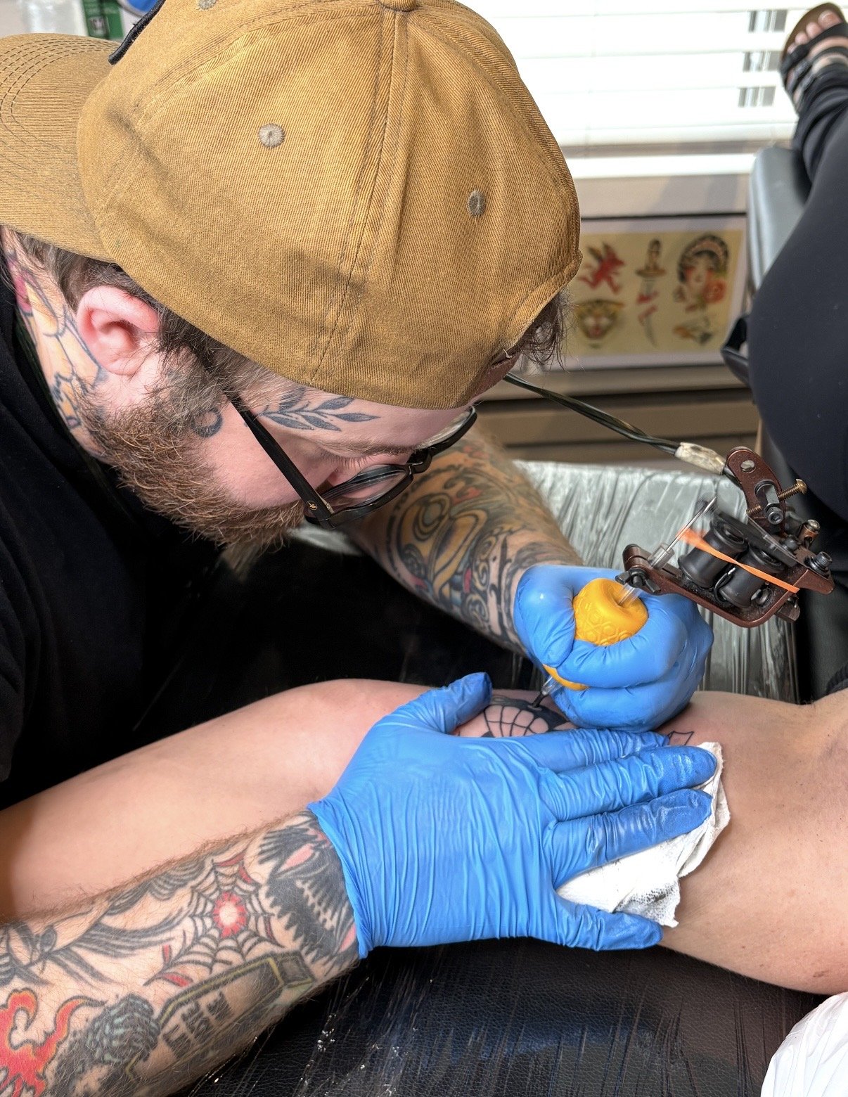 A tattooed man with tattoos on his arm and neck is tattooing on his clients leg. He is wearing a tan cap, glasses, and blue gloves. The tattoo artist is using a tattoo machine and a yellow grip, focusing on the tattoo work.