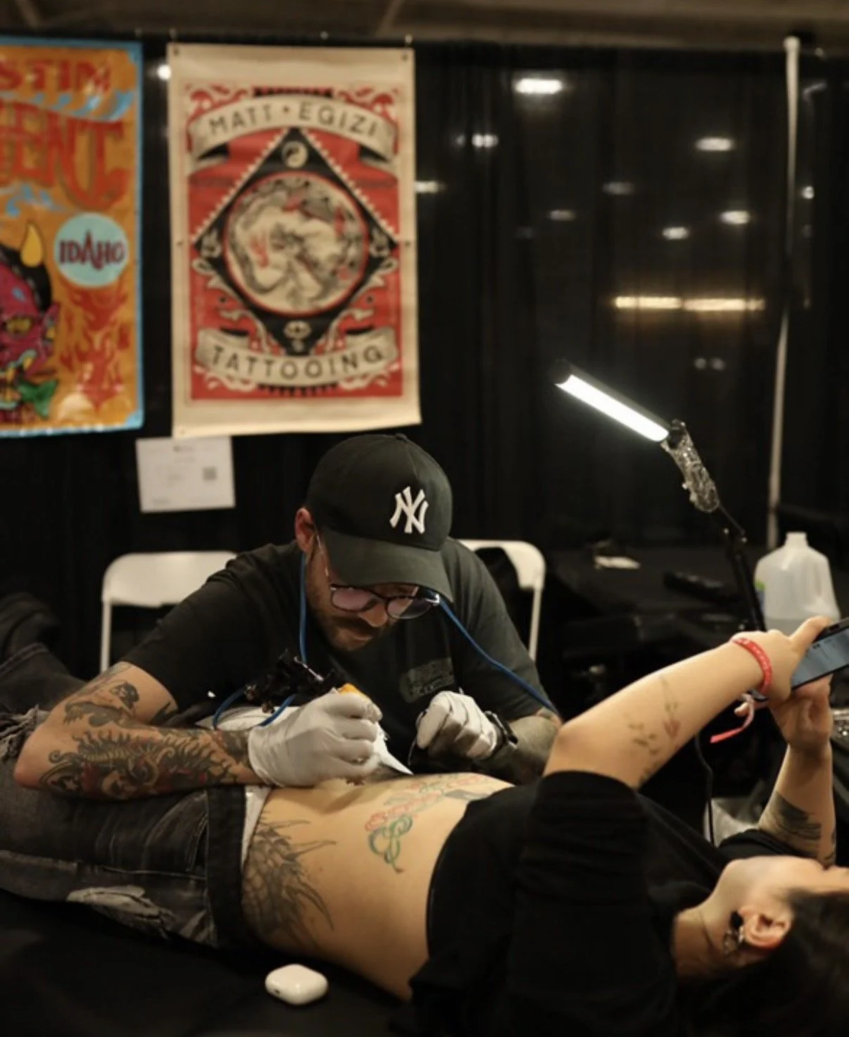 Tattoo artist working on a person's torso during a tattoo session at a tattoo convention with tattoo art posters on the wall.