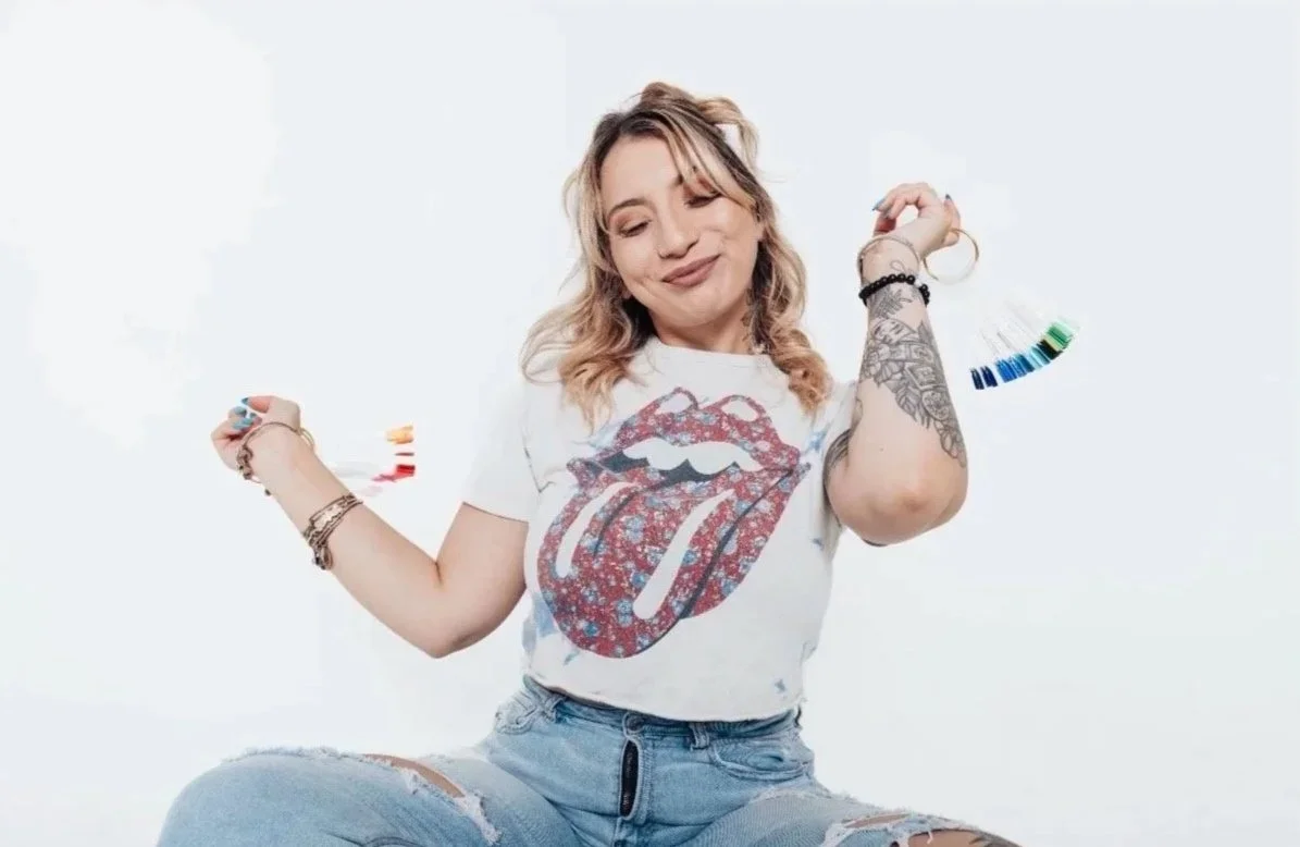 A young woman with blonde waves, wearing a Rolling Stones tongue logo t-shirt, ripped jeans, holding paint swatches, and smiling with eyes closed against a white background.