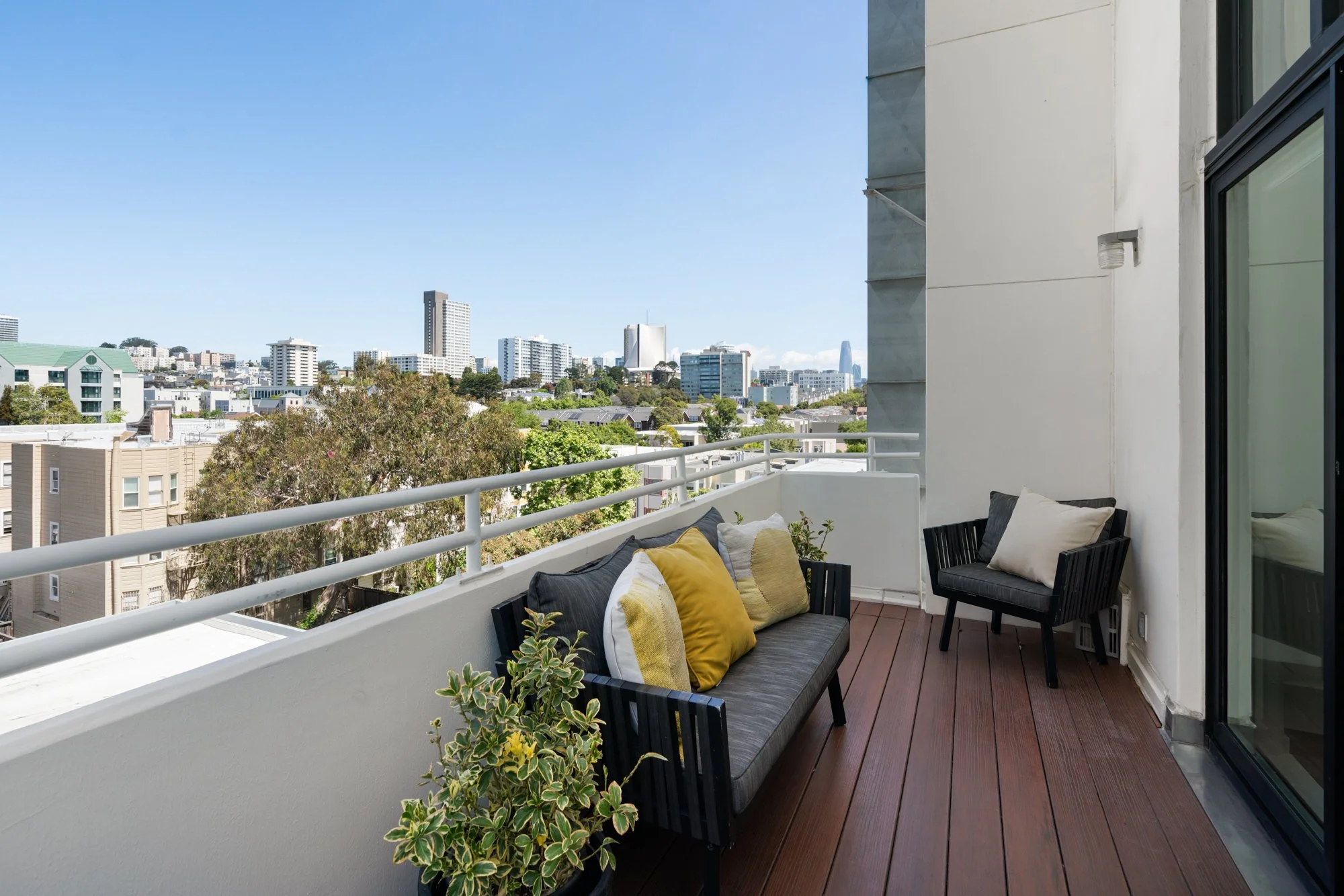 Balcony with sofa and chairs facing city skyline with tall buildings and trees under a clear blue sky.
