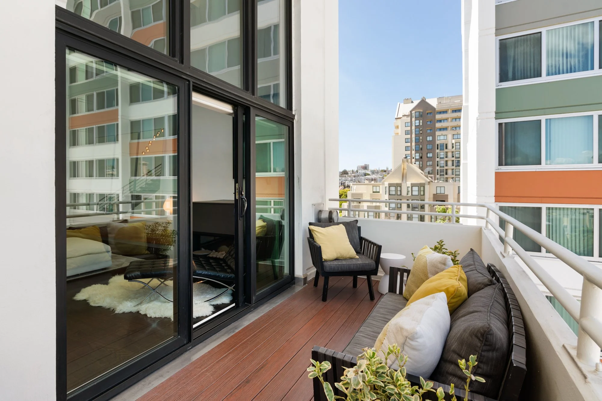 Balcony with outdoor seating, cushions, and potted plants overlooking adjacent modern apartment buildings under a clear blue sky.