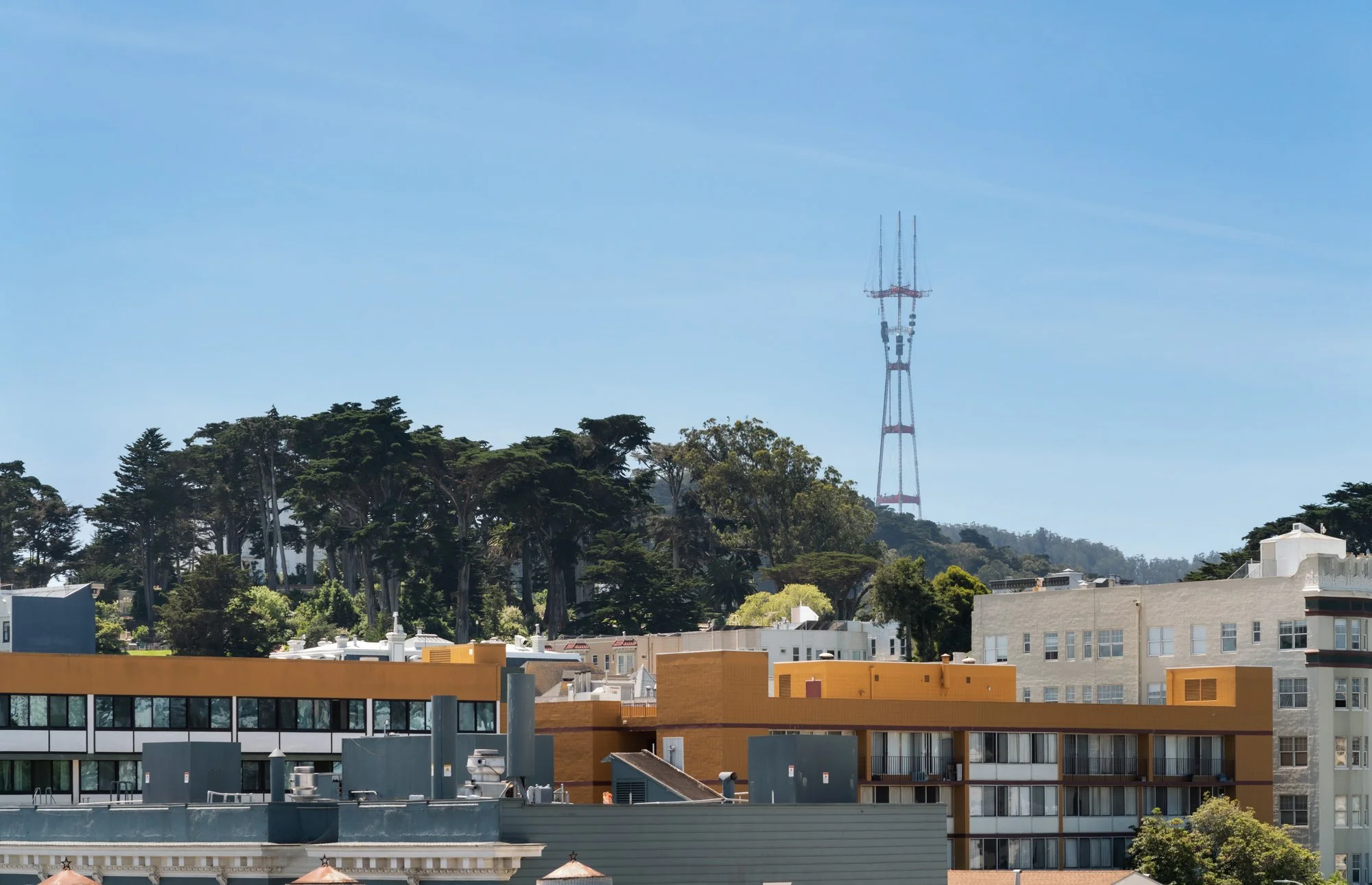 Cityscape with modern buildings in the foreground, tall trees in the middle, and a large red and white radio tower on a hill in the background under a clear blue sky.