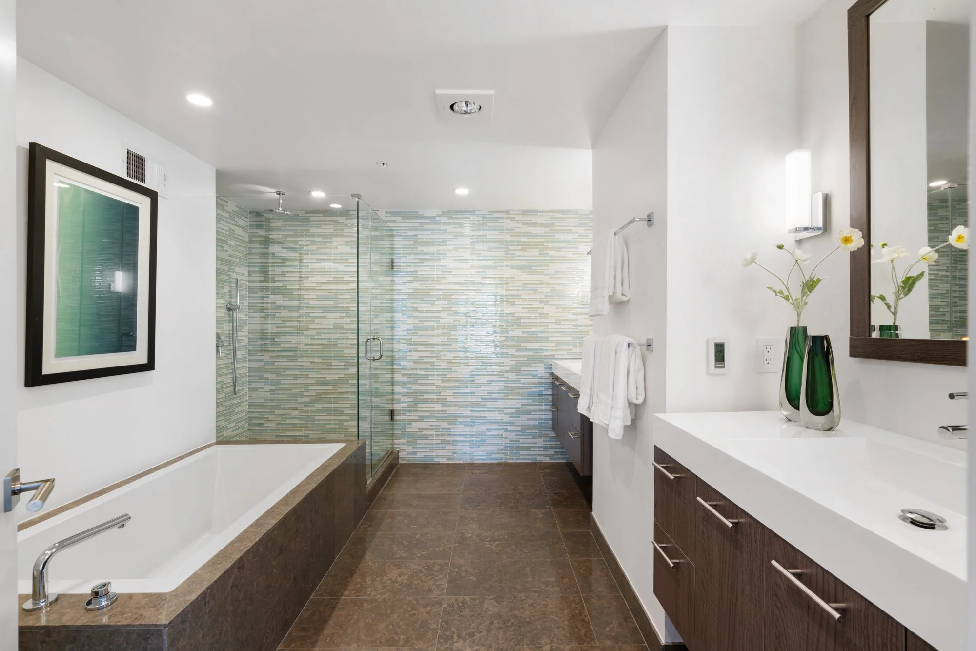 Modern bathroom with white walls, a glass-enclosed shower with multicolored tile, a bathtub, dark wood cabinetry, and decorative vases with flowers.