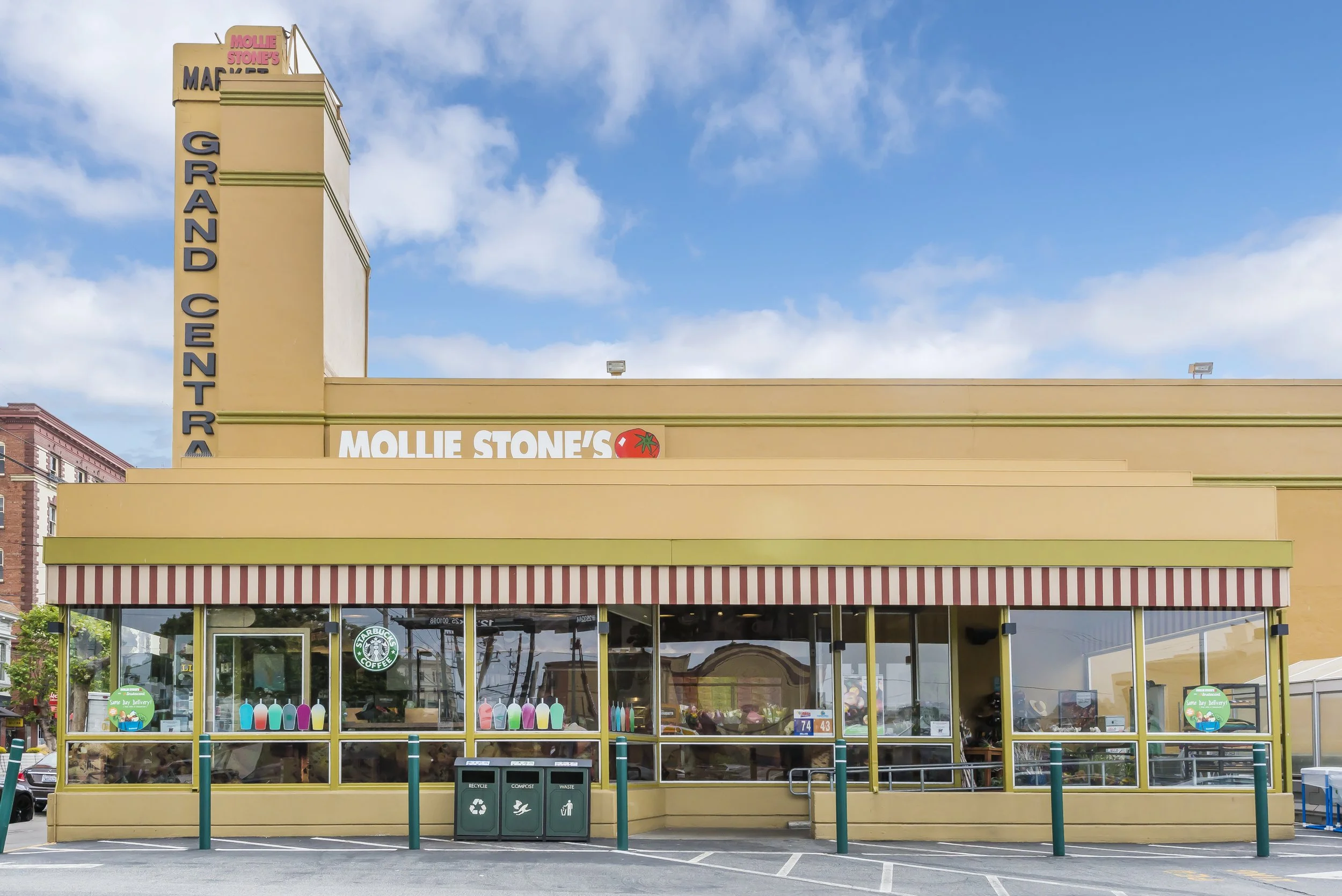 Exterior of a retail store with a yellow facade and red and white striped awning, large glass windows, and signage for Mollie Stone's grocery store. There is a parking lot in front with green poles, garbage bins, and a Starbucks logo visible inside t