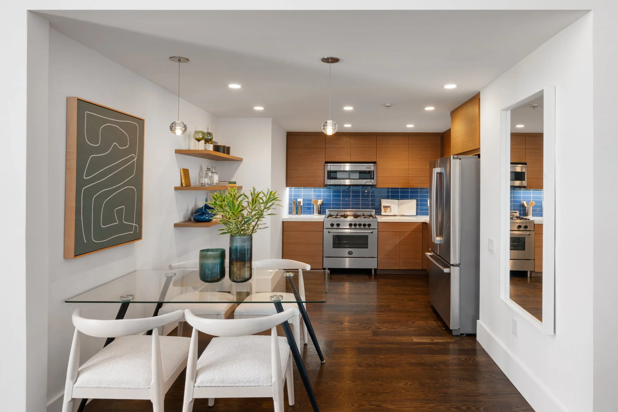 Modern kitchen with wooden cabinets, blue tile backsplash, stainless steel appliances, and a dining area featuring a glass table with white chairs and decorative vases.