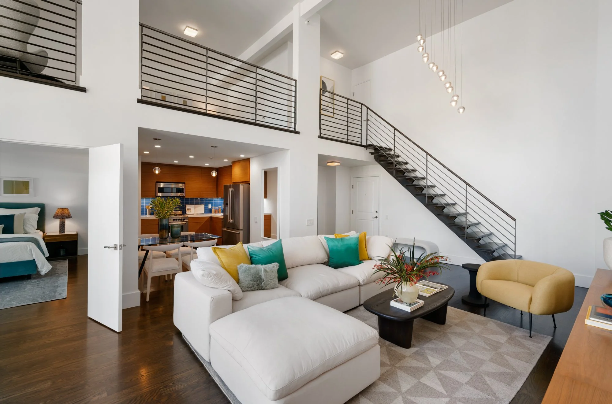 Interior view of a modern, spacious living area with white walls, wooden floors, a white sectional sofa with colorful pillows, a dark wood coffee table, and a yellow armchair. A staircase with dark rails leads to an upper level, and there is a view o