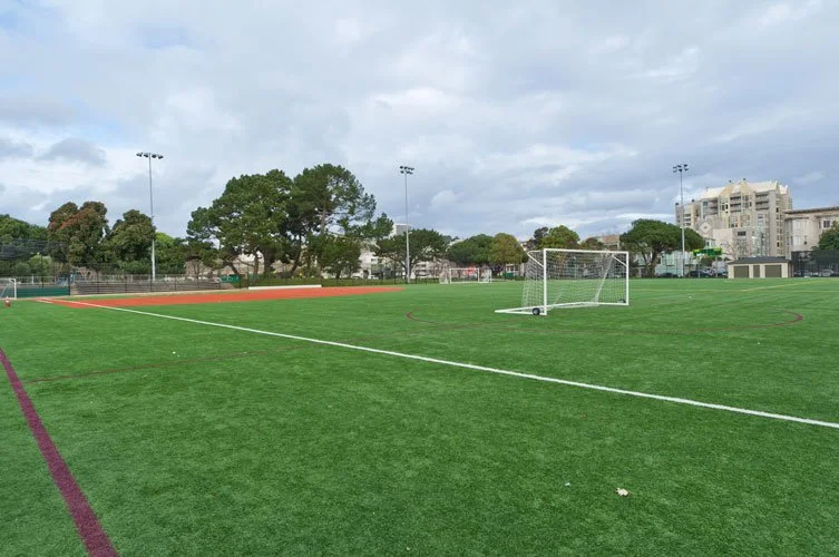An empty sports field with artificial turf, a soccer goal, and a running track, surrounded by trees and buildings under a cloudy sky.