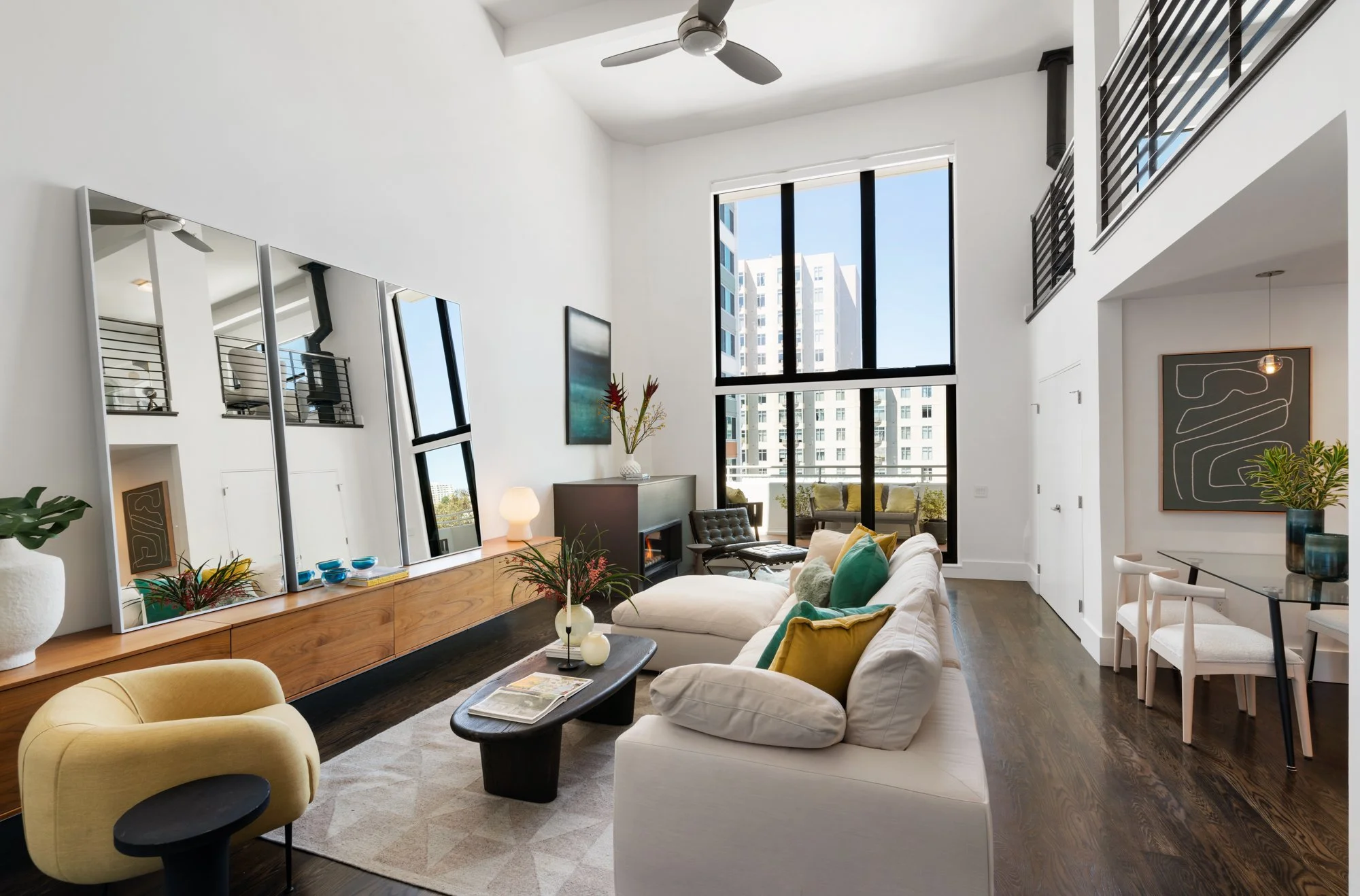 Living room with a large window, white walls, modern furniture including a white sofa with colorful pillows, a yellow armchair, a black coffee table, a long wooden sideboard with three mirrors, a TV, and decorative plants. There is a high ceiling wit
