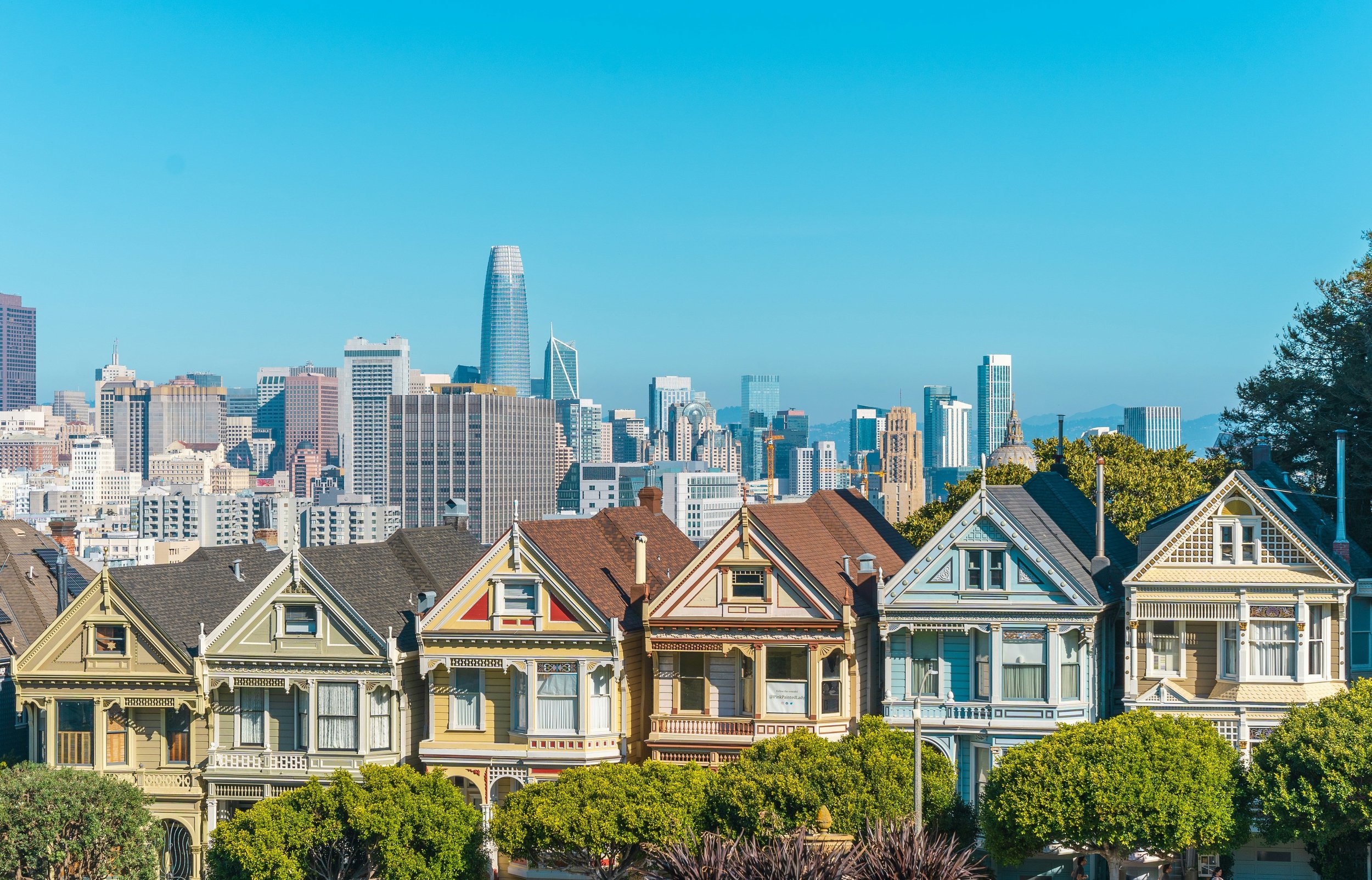 A view of San Francisco skyline with Victorian houses in the foreground under a clear blue sky.