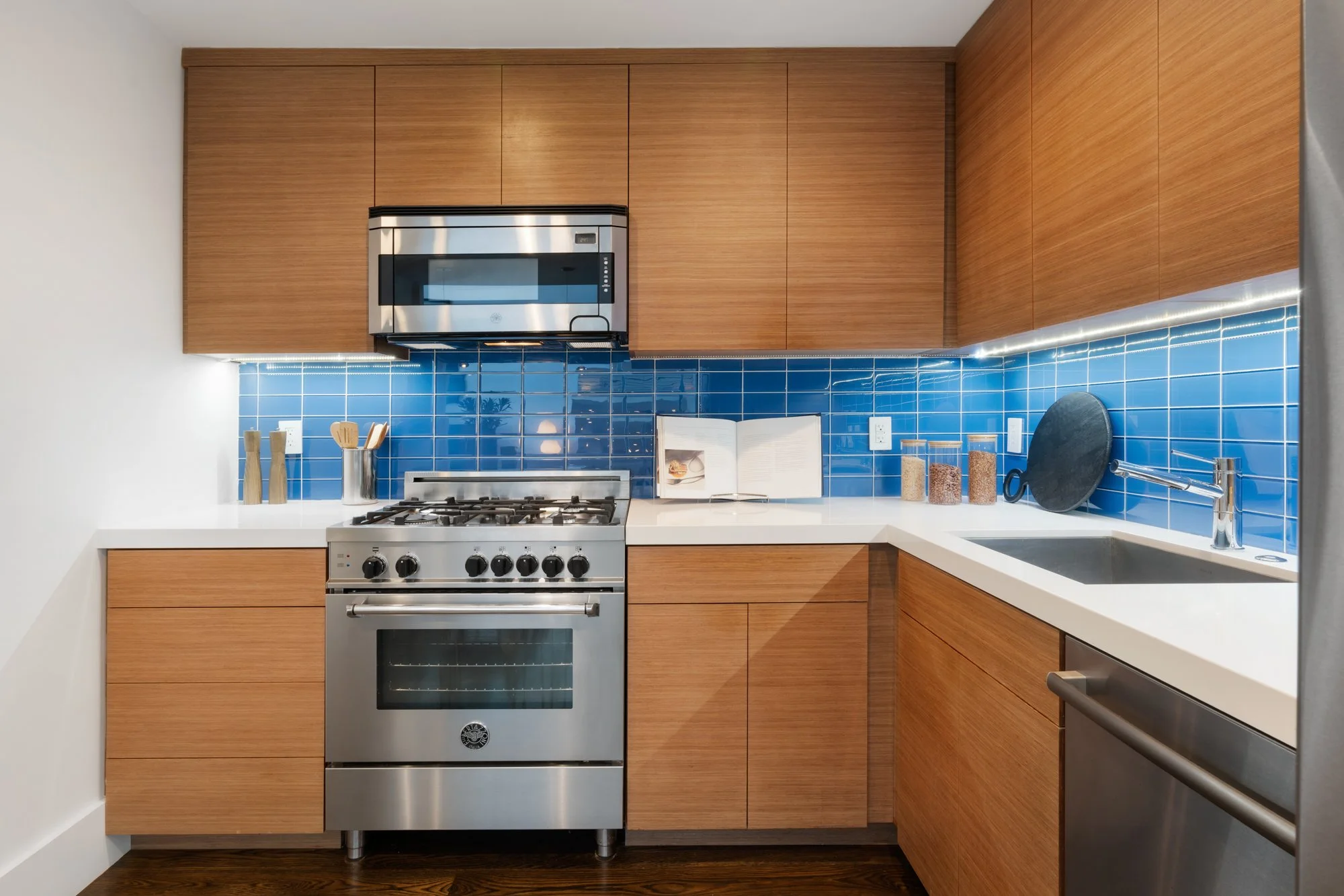 Modern kitchen with wooden cabinets, a stainless steel oven, a blue tiled backsplash, and various kitchen utensils and containers on the white countertop.