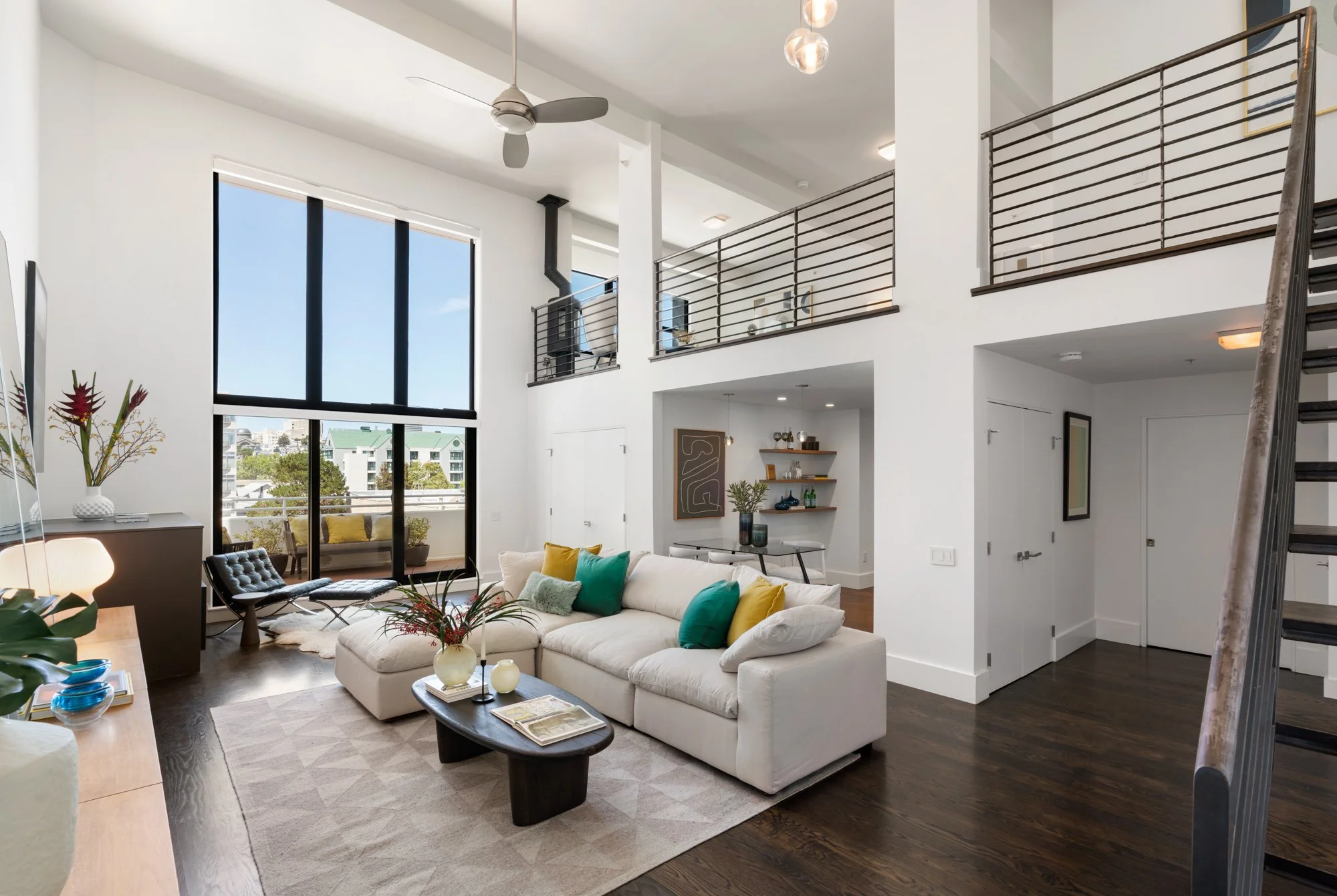 Modern open-concept living room with large windows, white walls, a vaulted ceiling with ceiling fan, colorful throw pillows on a white sectional sofa, contemporary decor, and a balcony view of neighboring buildings.