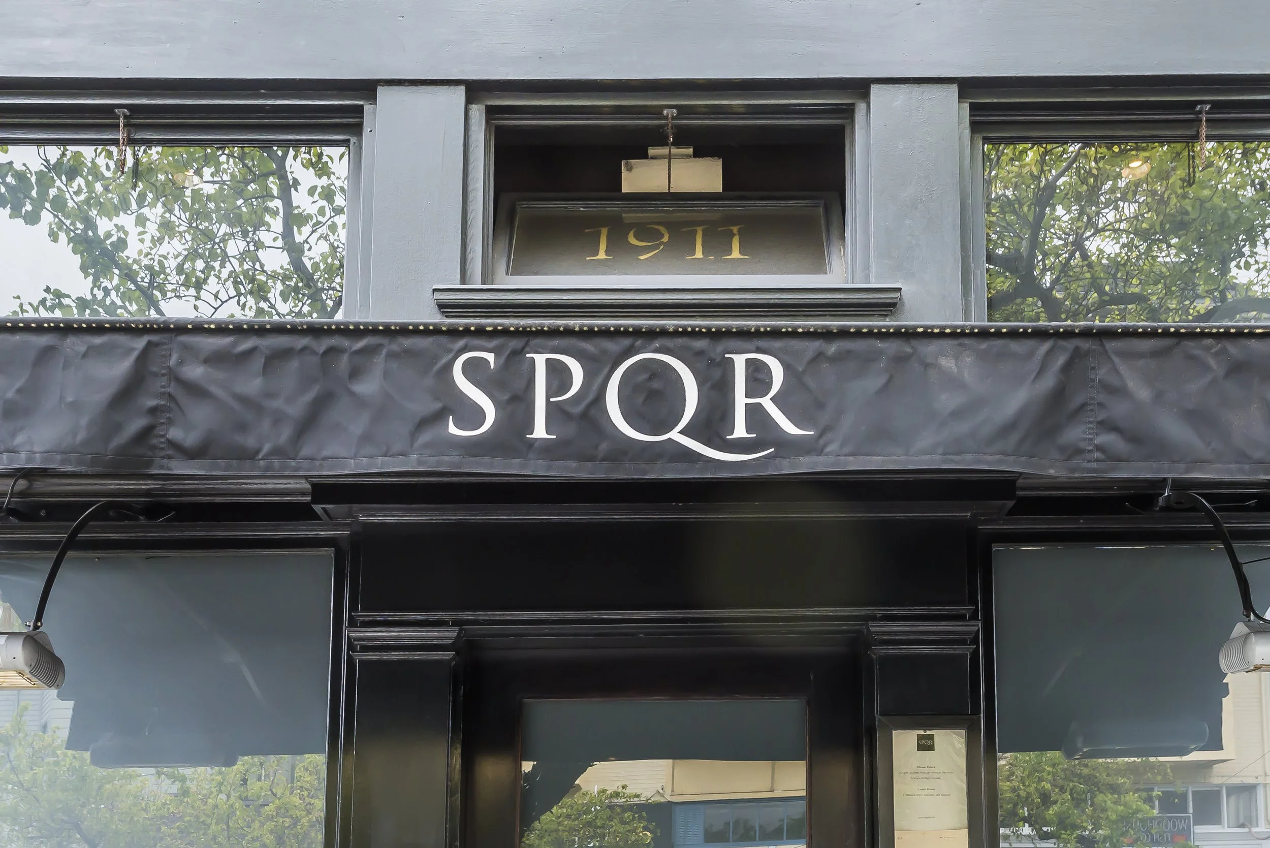 The image shows a building facade with two large windows reflecting trees, and a sign with the text 'SPORT' hanging below a ledge. The middle section displays a bronze or gold-colored sign with the numbers '1911'.