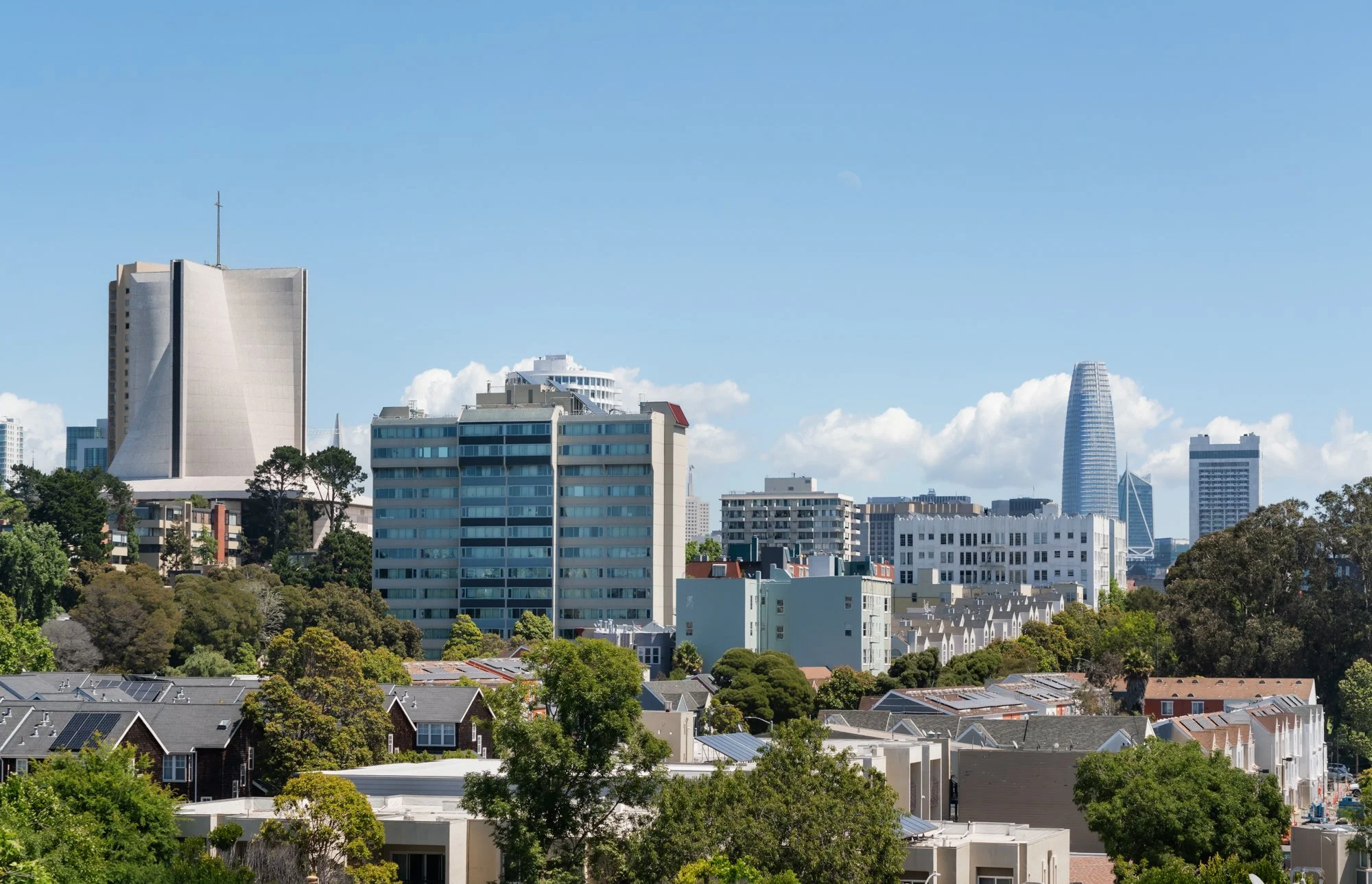 A city skyline with tall buildings, including a distinctive tapered skyscraper, under a blue sky with white clouds, with trees and residential homes in the foreground.