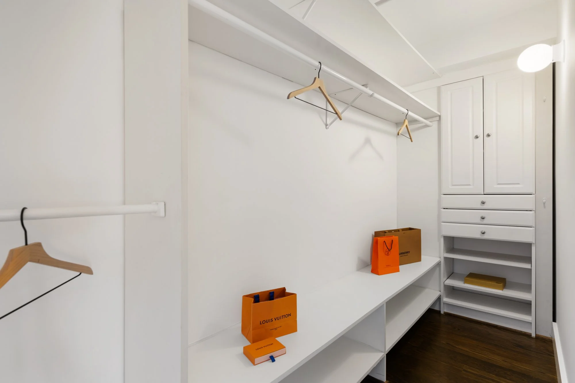 Empty walk-in closet with white shelves, drawers, and hanging rods, some with beige hangers, and Louis Vuitton shopping bags on the shelves.