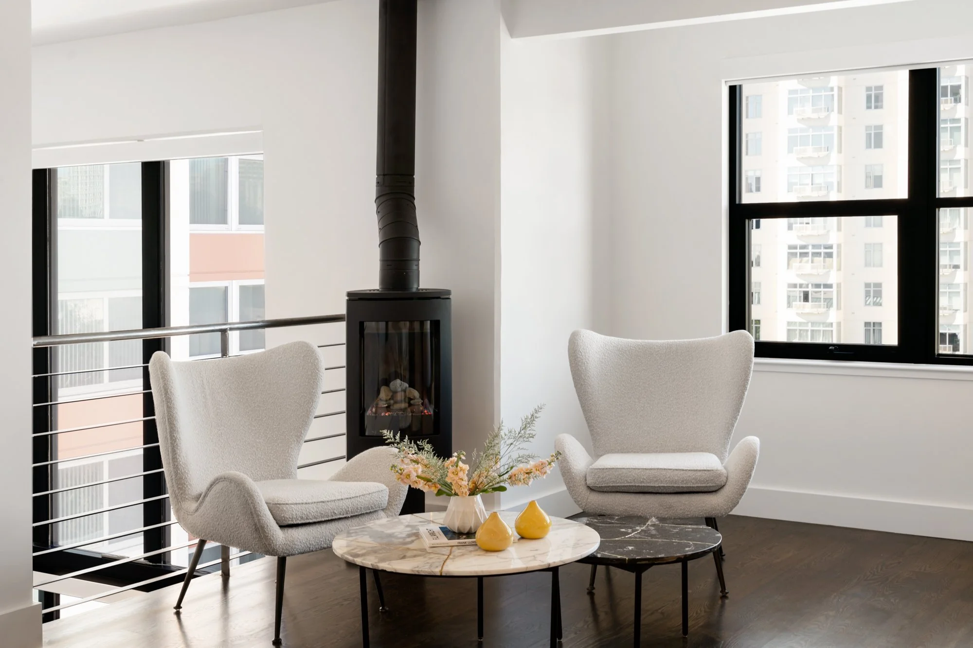 Modern living room with two white upholstered armchairs, a marble coffee table with a small bouquet of pink flowers in a white vase, and three yellow decorative vases. Large windows with black frames let in natural light, and a black wood stove with 