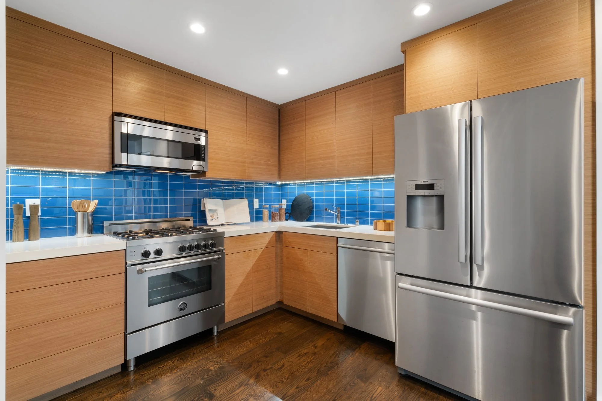 Modern kitchen with wooden cabinets, stainless steel appliances including a refrigerator, microwave, and gas stove, blue tiled backsplash, dark wood flooring, and recessed ceiling lights.