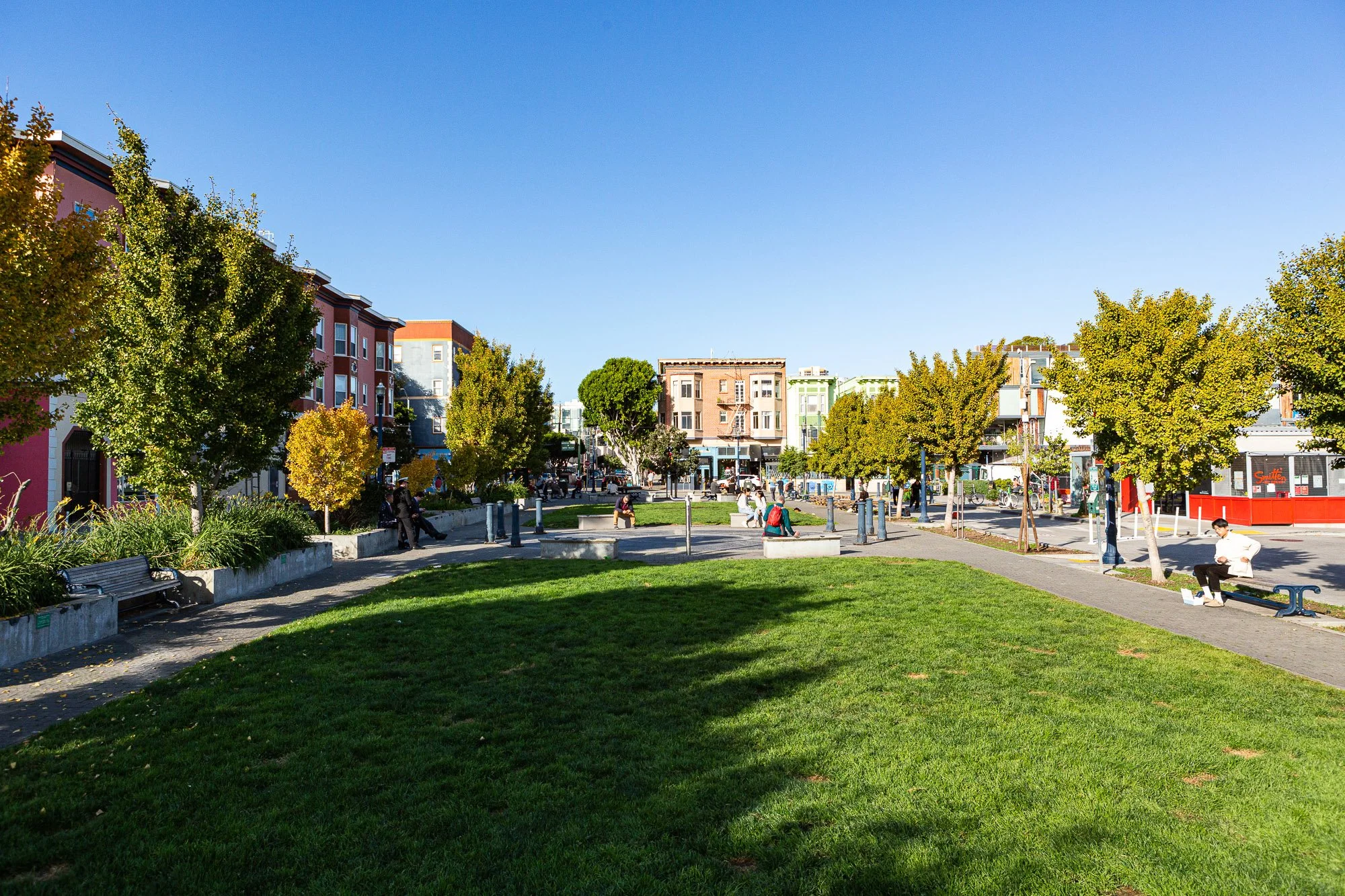 A vibrant urban park with green grass and trees, surrounded by colorful buildings. People are sitting on benches and walking, enjoying a sunny day in the city.