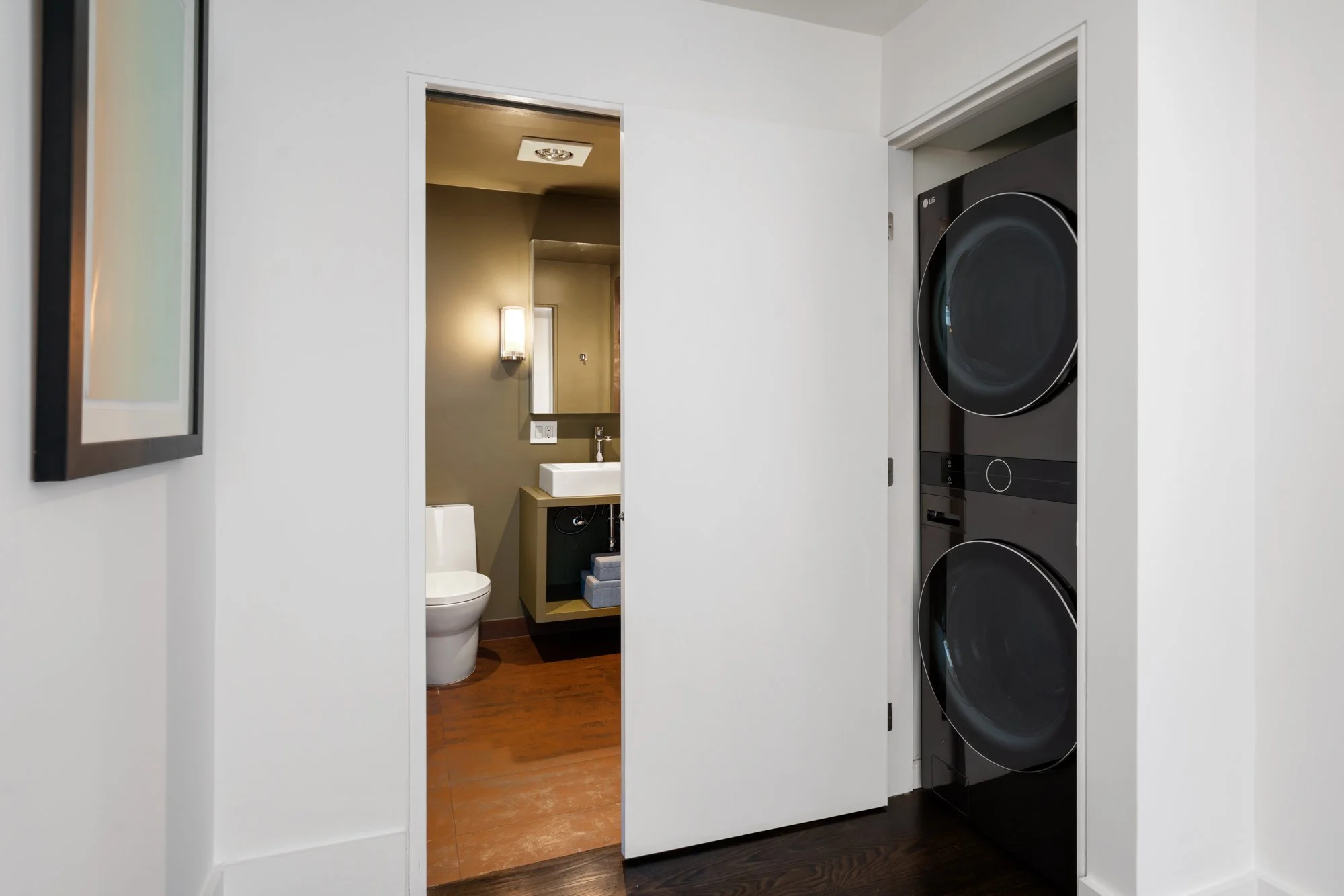 Laundry area with stacked washing machine and dryer on the right, view into a small bathroom with toilet, sink, and beige walls.