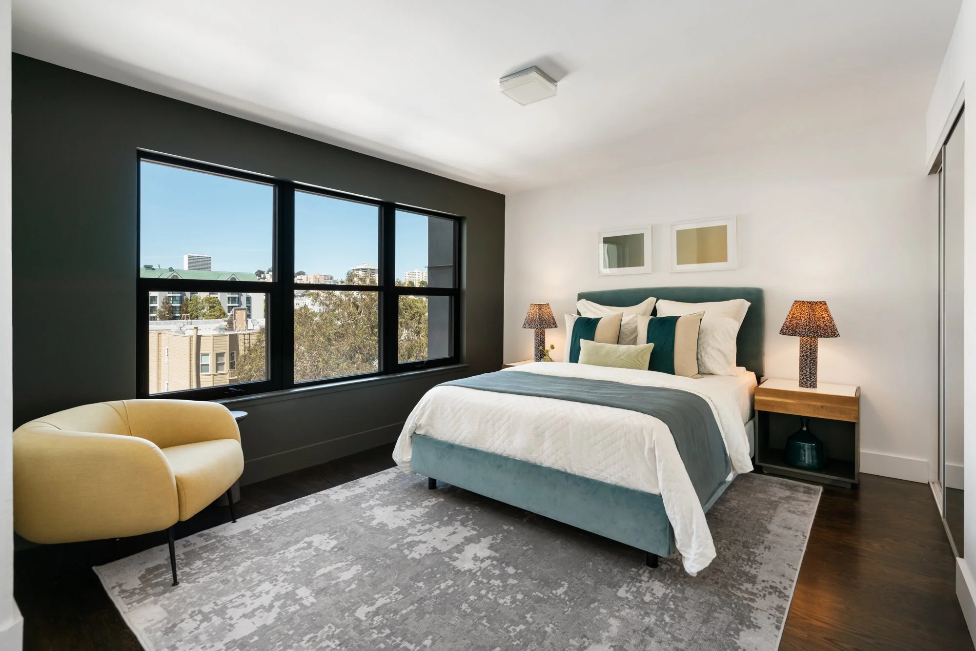 Modern bedroom with large window, dark accent wall, bed with teal headboard, and beige and white bedding. Two bedside tables with matching lamps, and a yellow armchair in the corner.