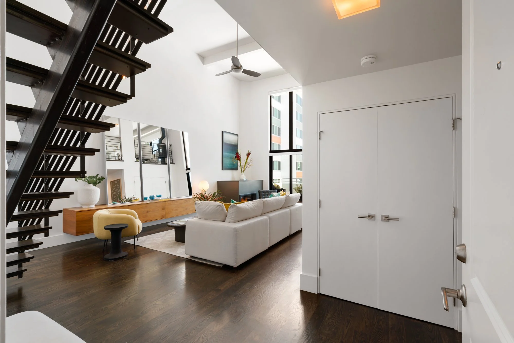Modern living room with white sofa, wooden sideboard, plant, art, mirrors, large windows, and stairs.