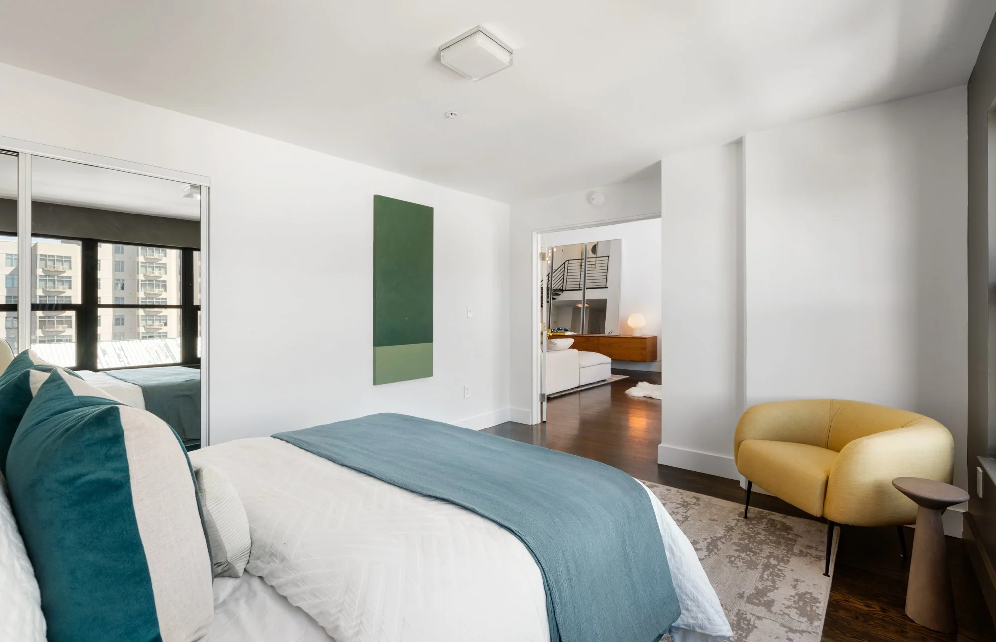 Modern living room with white walls, a bed with teal and white bedding, a yellow armchair, and a green abstract art piece.