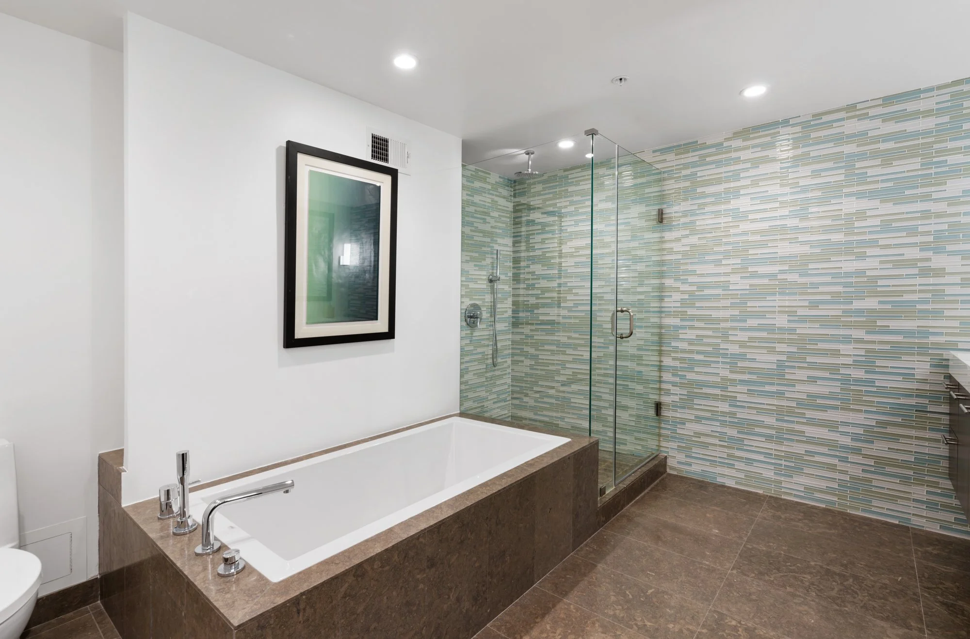 Modern bathroom with a bathtub, glass-enclosed shower, and a framed mirror on a white wall. The shower wall has a multicolored striped tile pattern, and the bathtub surround is finished with dark brown stone.