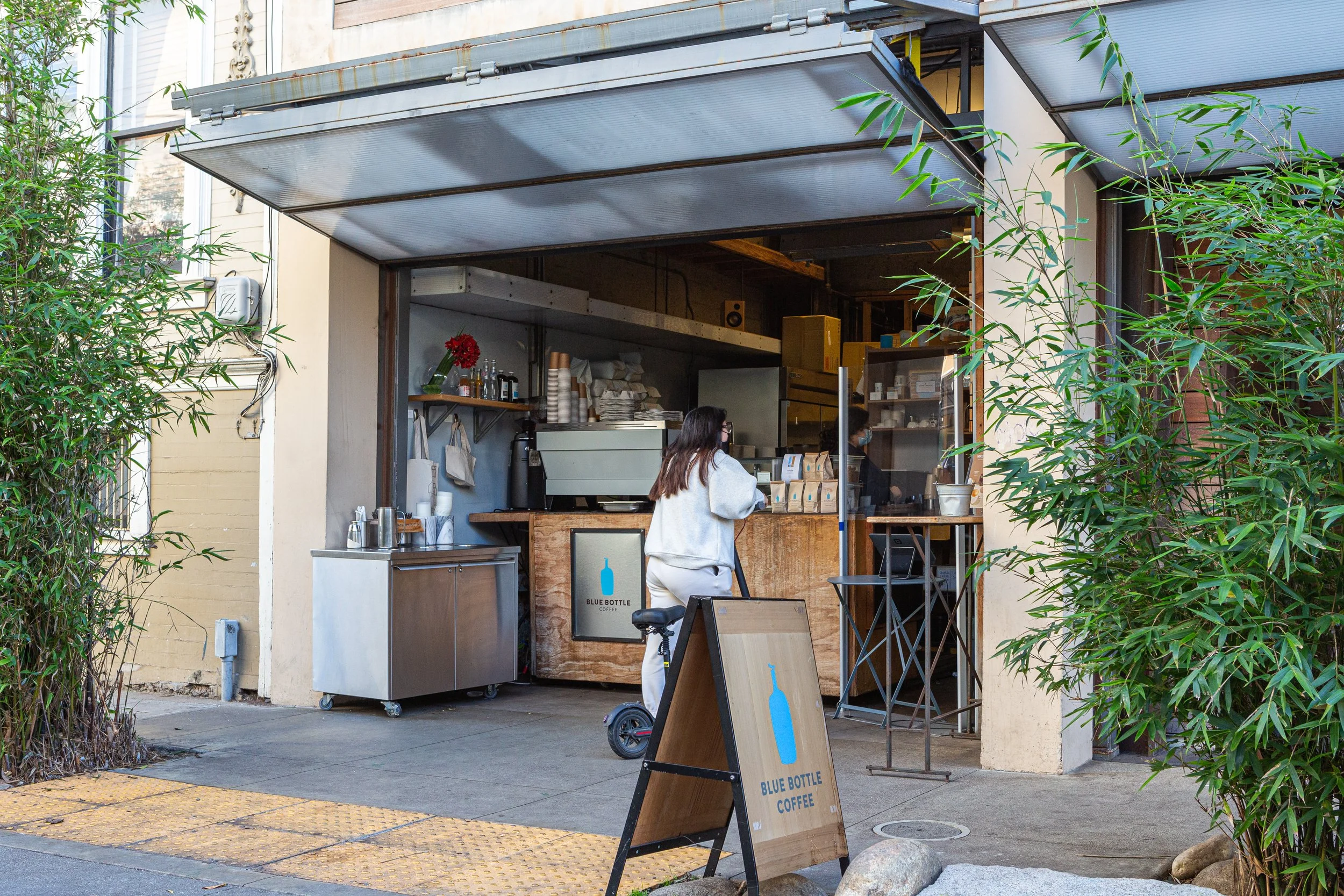 Outdoor coffee stand named Blue Bottle Coffee with a woman ordering, located on a sidewalk with trees and buildings around.
