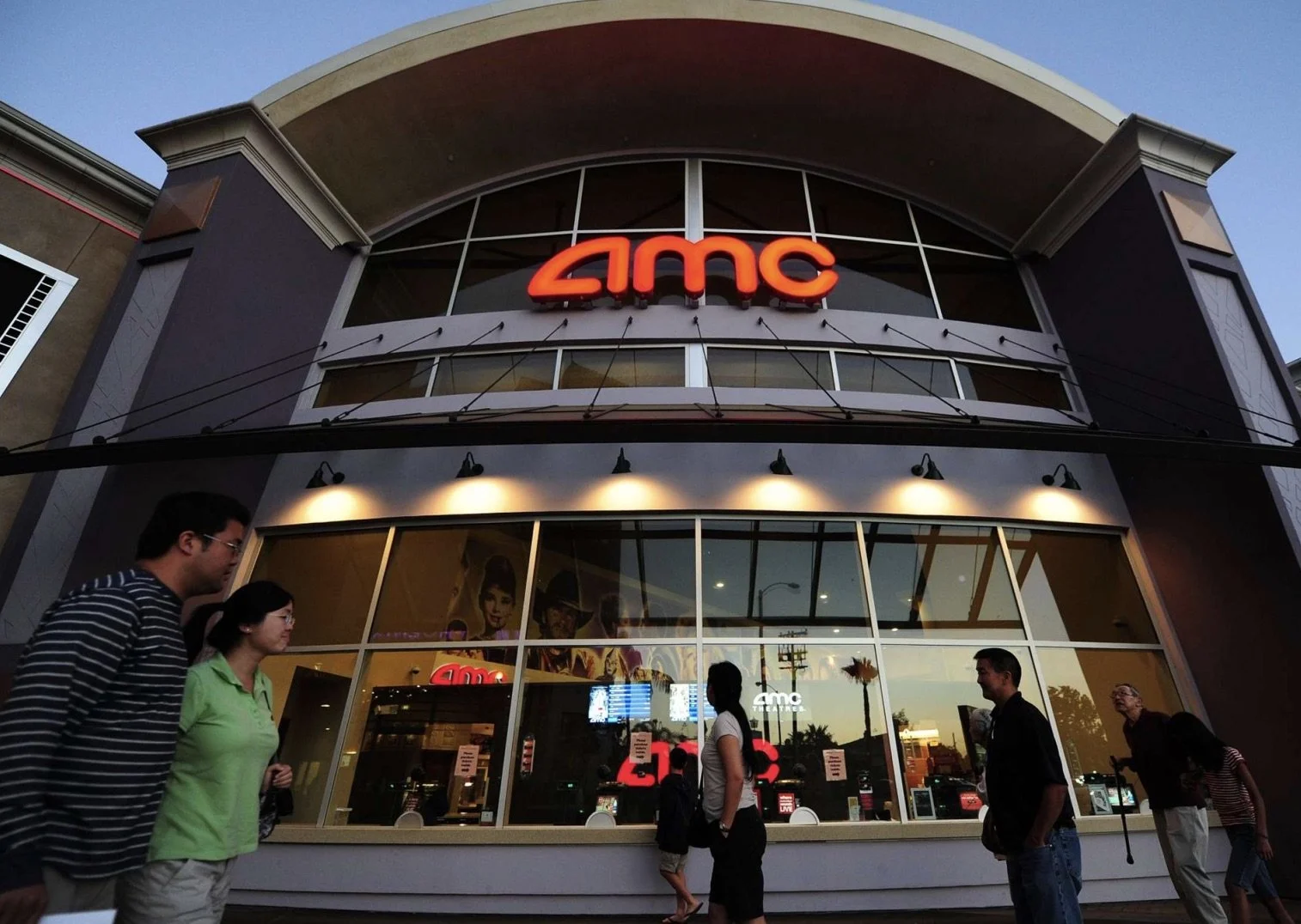 People walking past an AMC movie theater during dusk, with the AMC logo illuminated on the building facade and posters visible inside through the large glass windows.