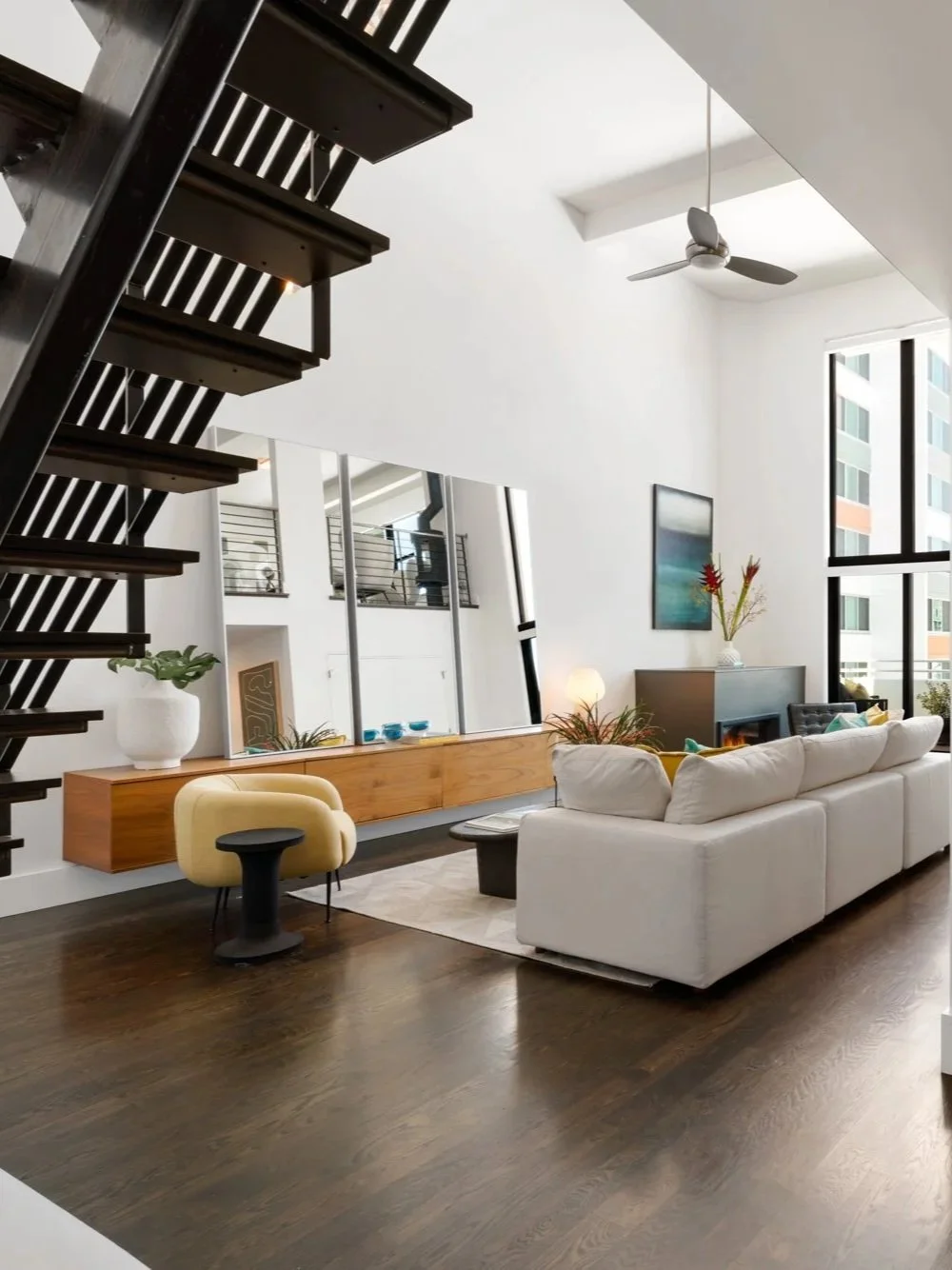 Modern living room with dark wooden flooring, white walls, large windows, a white sofa, a yellow chair, a black coffee table, a wall-mounted mirror, a mounted television, and decorative plants.