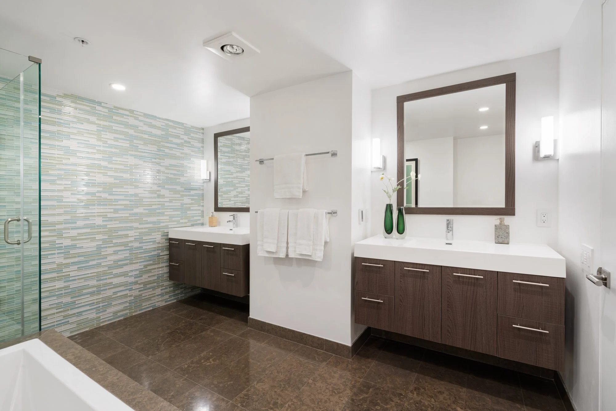 Modern bathroom with a glass-enclosed shower, double vanity with two sinks, large mirror, wooden cabinets, and white walls with decorative lighting and vases.