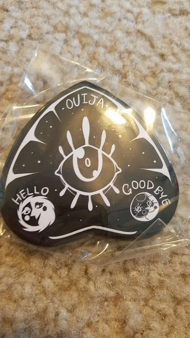 A black sticker with a doodle of a smiling eye with eyelashes, surrounded by words "OUJSA," "HELLO," and "GOODBYE."