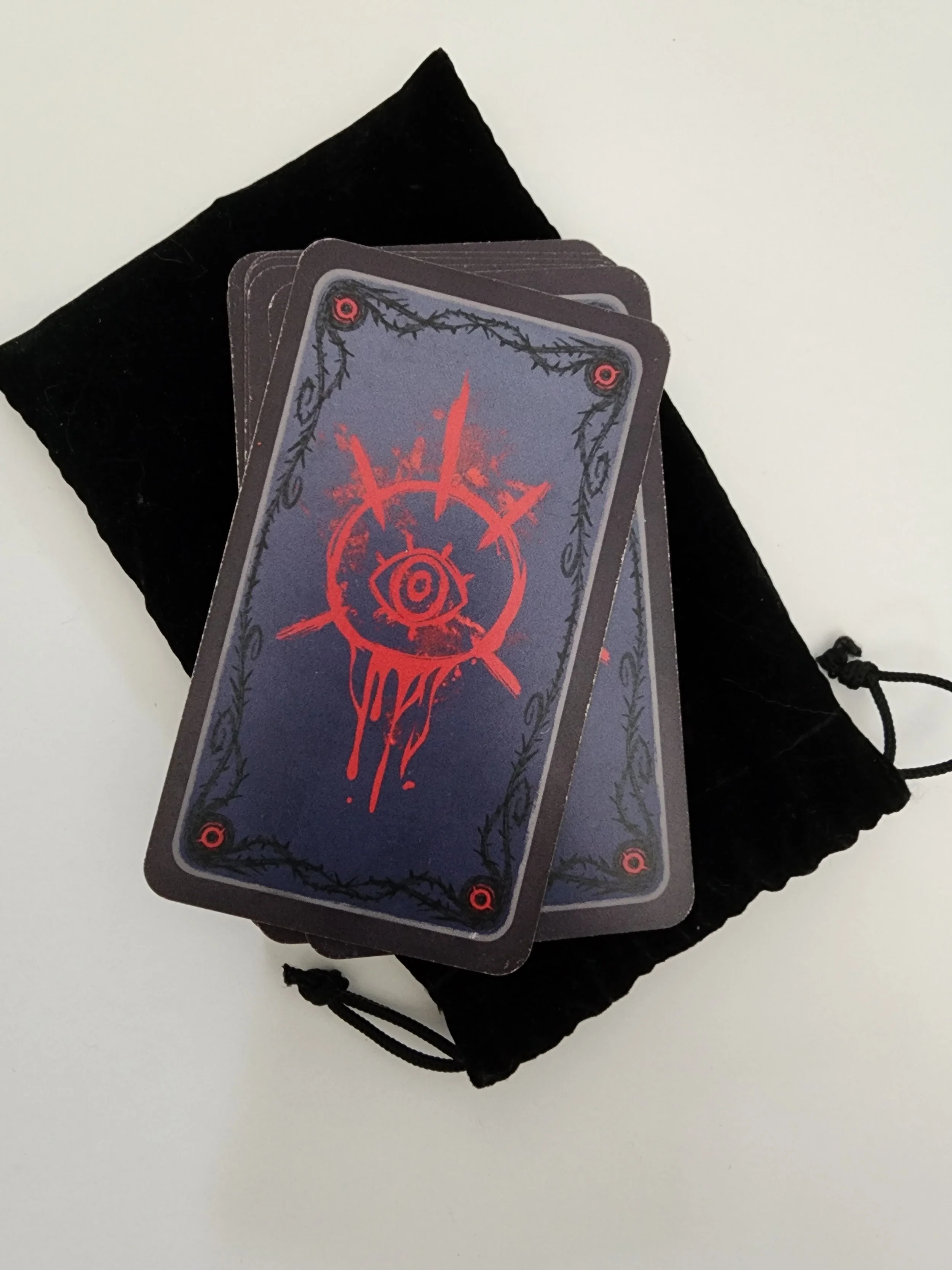 A deck of Tarot cards with a dark design, featuring a red symbol and black border, placed on a black velvet bag with a drawstring, on a white surface.