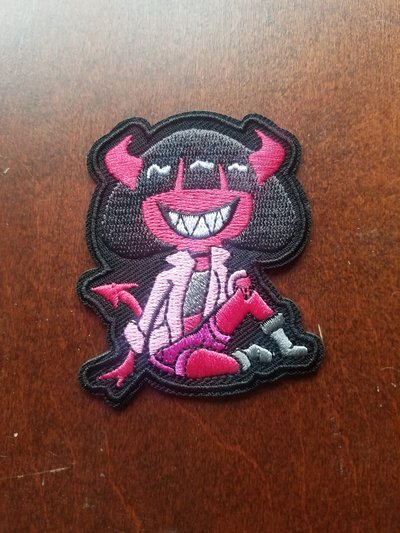 A cartoon patch of a smiling creature with pink horns and a fang-filled grin, wearing a light-colored shirt and pink pants, sitting on a surface.