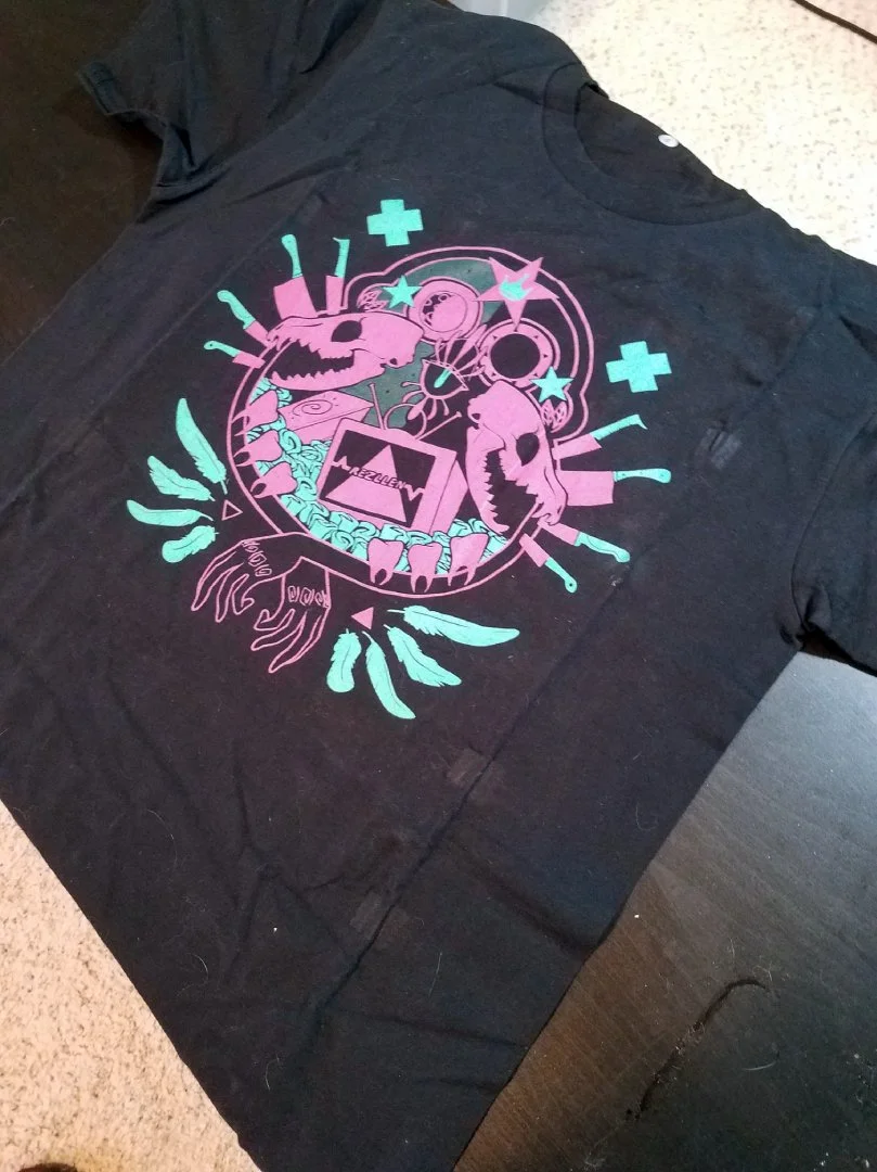 Black T-shirt with a colorful graphic design featuring skulls, feathers, stars, and abstract shapes in pink, blue, and purple.