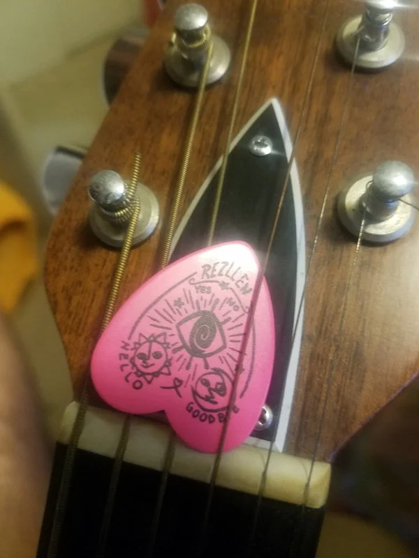 Close-up of an electric guitar headstock with tuning pegs and strings, featuring a pink guitar pick attached to the strings, decorated with handwritten doodles and text.