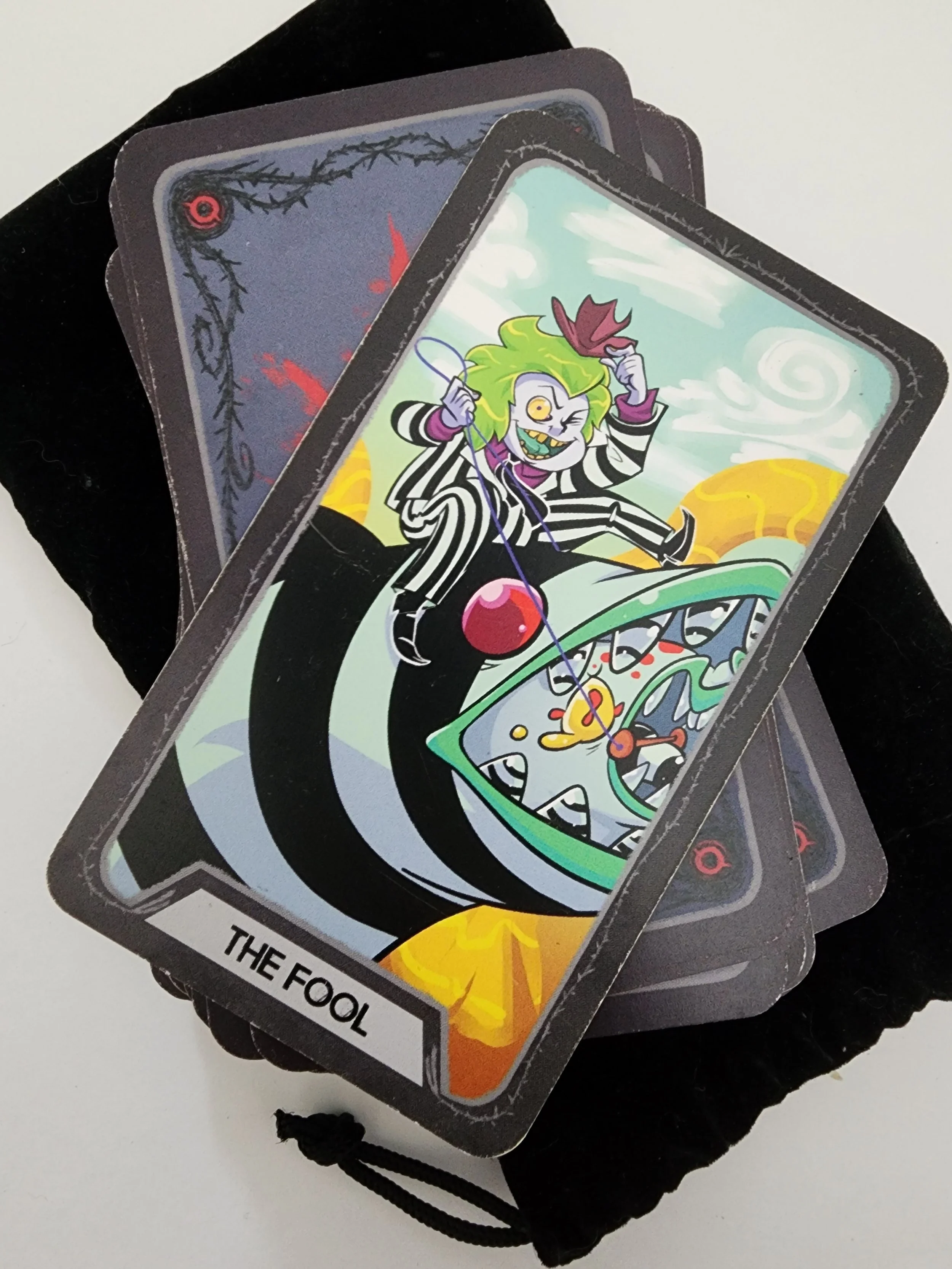 A deck of tarot cards featuring a cartoon-style illustration of a joker character with green hair, a winking eye, and striped clothing, holding a purple fan, with background elements including clouds, a sun, and swirling patterns.