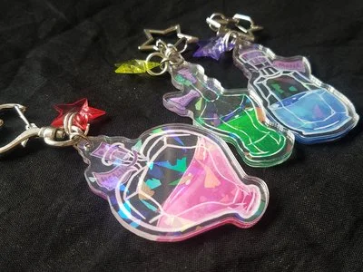Three acrylic keychains shaped like cat heads with holographic designs, attached to metal clips with colored stars, placed on a black surface.