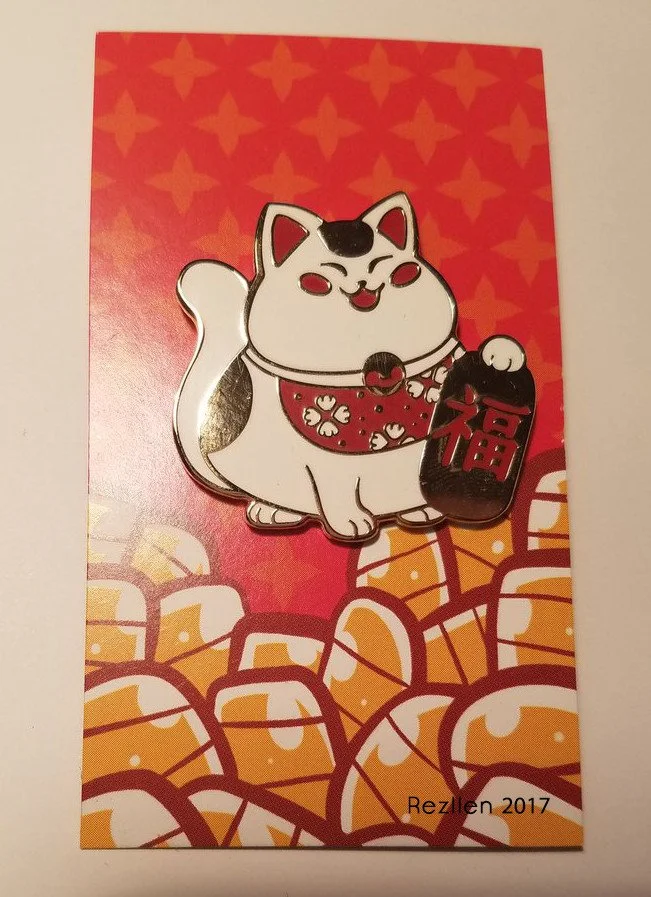 A pin of a cheerful white maneki-neko with a black spot on its head, holding a traditional object with the Chinese character for good fortune (福). The background has a red and gold pattern with stylized coins.
