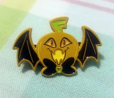 A Halloween-themed pin featuring a smiling pumpkin with bat wings, a green leaf on top, and a small green spider at the bottom.