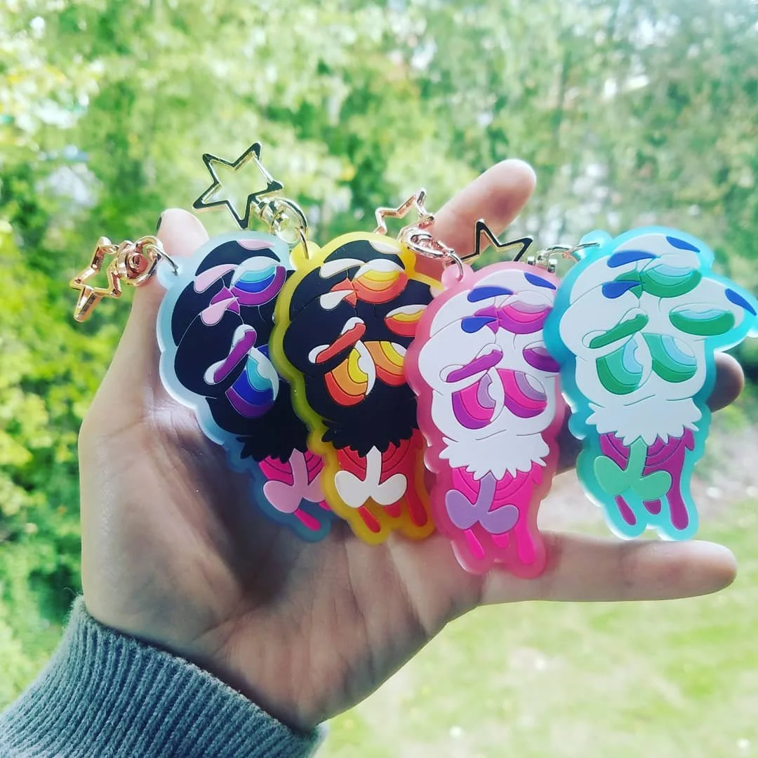Colorful acrylic keychains with cartoon character design, each featuring star-shaped metal attachments, held in a person's hand outdoors with green trees in the background.