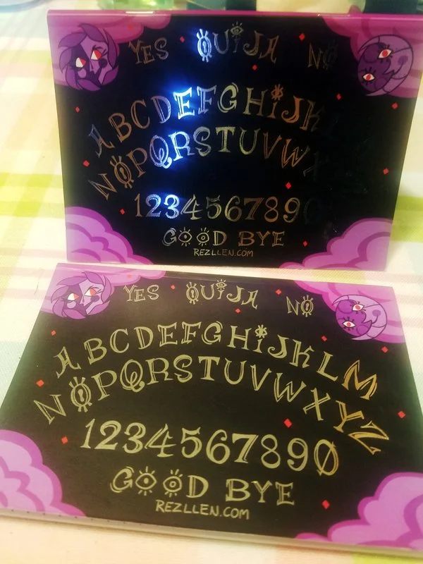 A black Ouija board with gold and purple lettering, featuring a purple cartoon character with eyes on the corners, and pink clouds at the bottom.