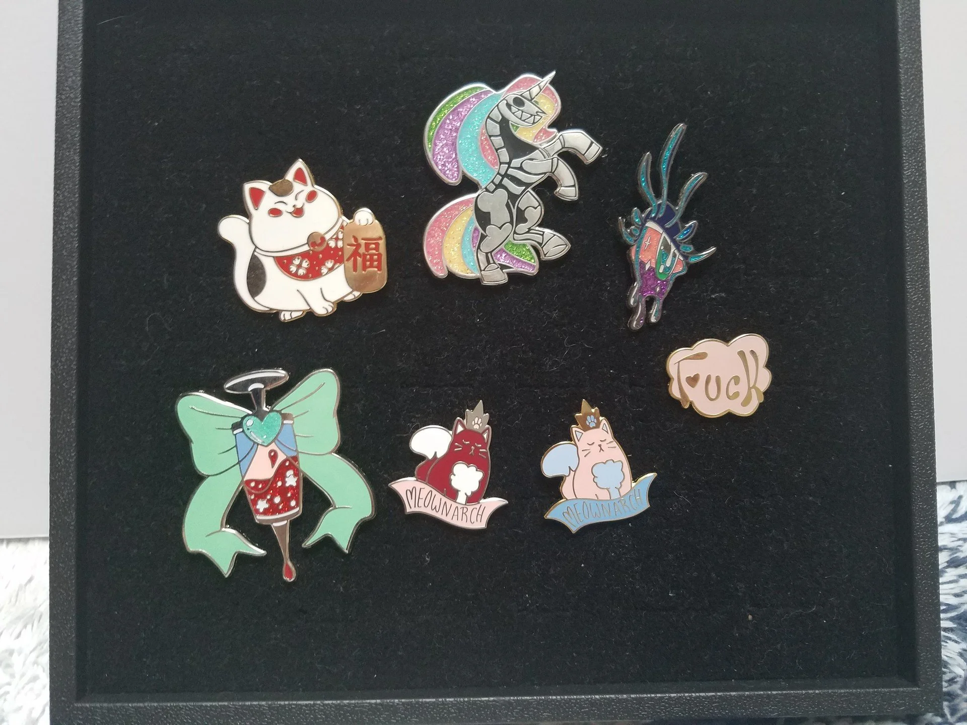 Collection of colorful enamel pins featuring various animals and characters, including a cat with a gold ingot, a unicorn with rainbow mane, a stylized deer, and cats with text reading 'Meow March' and 'Yuck', arranged on a black display backing.