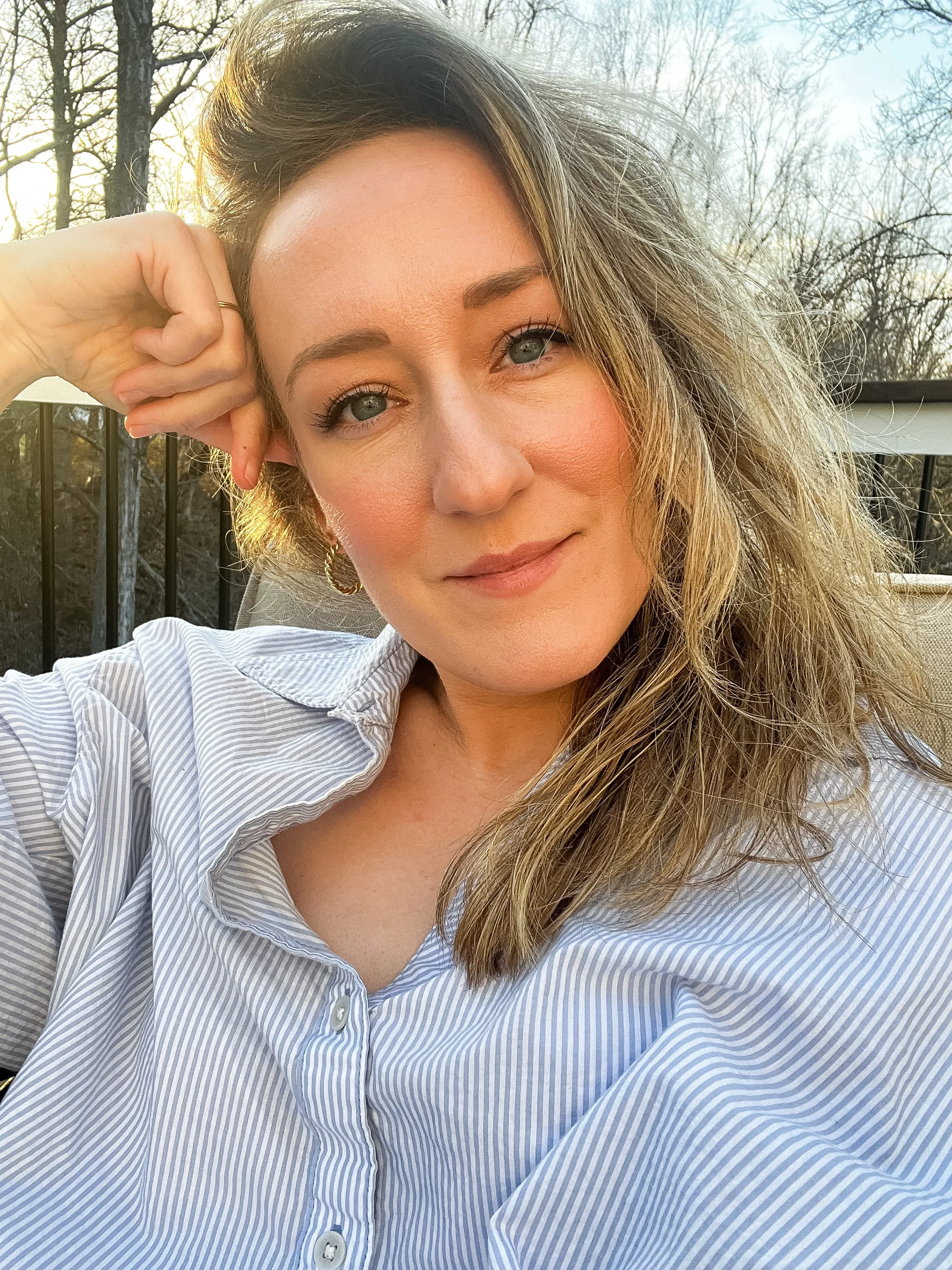 Close-up of a woman with wavy blonde hair, light skin, and blue eyes, wearing a striped button-up shirt and gold hoop earrings, sitting outdoors with a background of bare trees and a black railing.