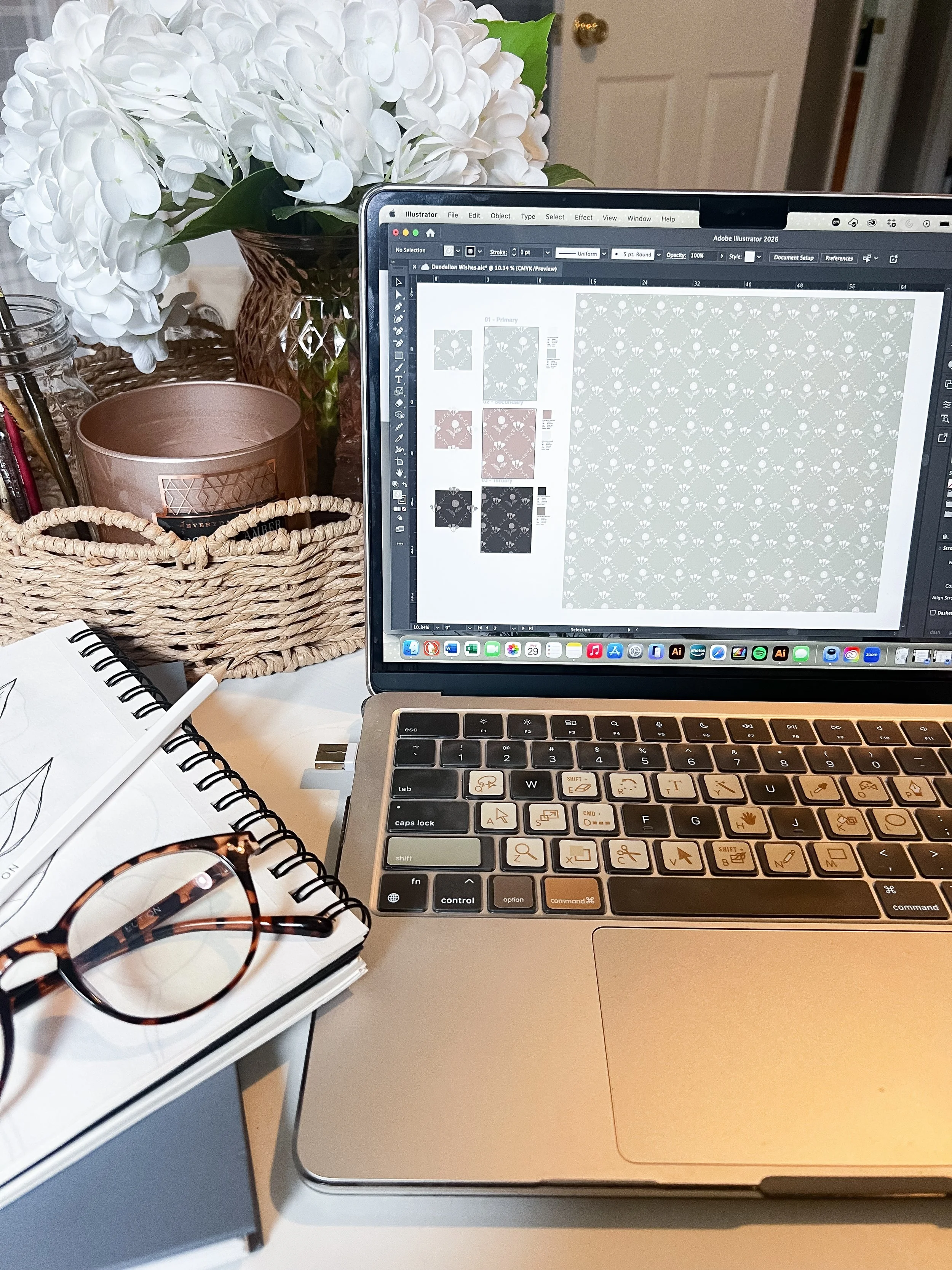 A workspace with a MacBook laptop displaying Adobe Illustrator on the screen, a spiral notebook with a pen and sunglasses resting on top, a basket, a pink candle holder, and a large bouquet of white hydrangeas in a glass vase.
