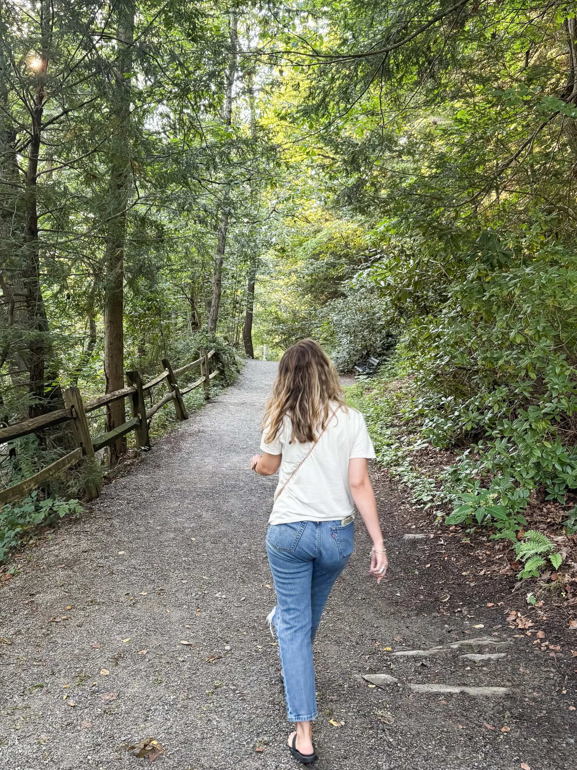 A woman with wavy hair, wearing a white t-shirt, blue jeans, and black slide sandals, walking on a gravel path through a lush green forest.